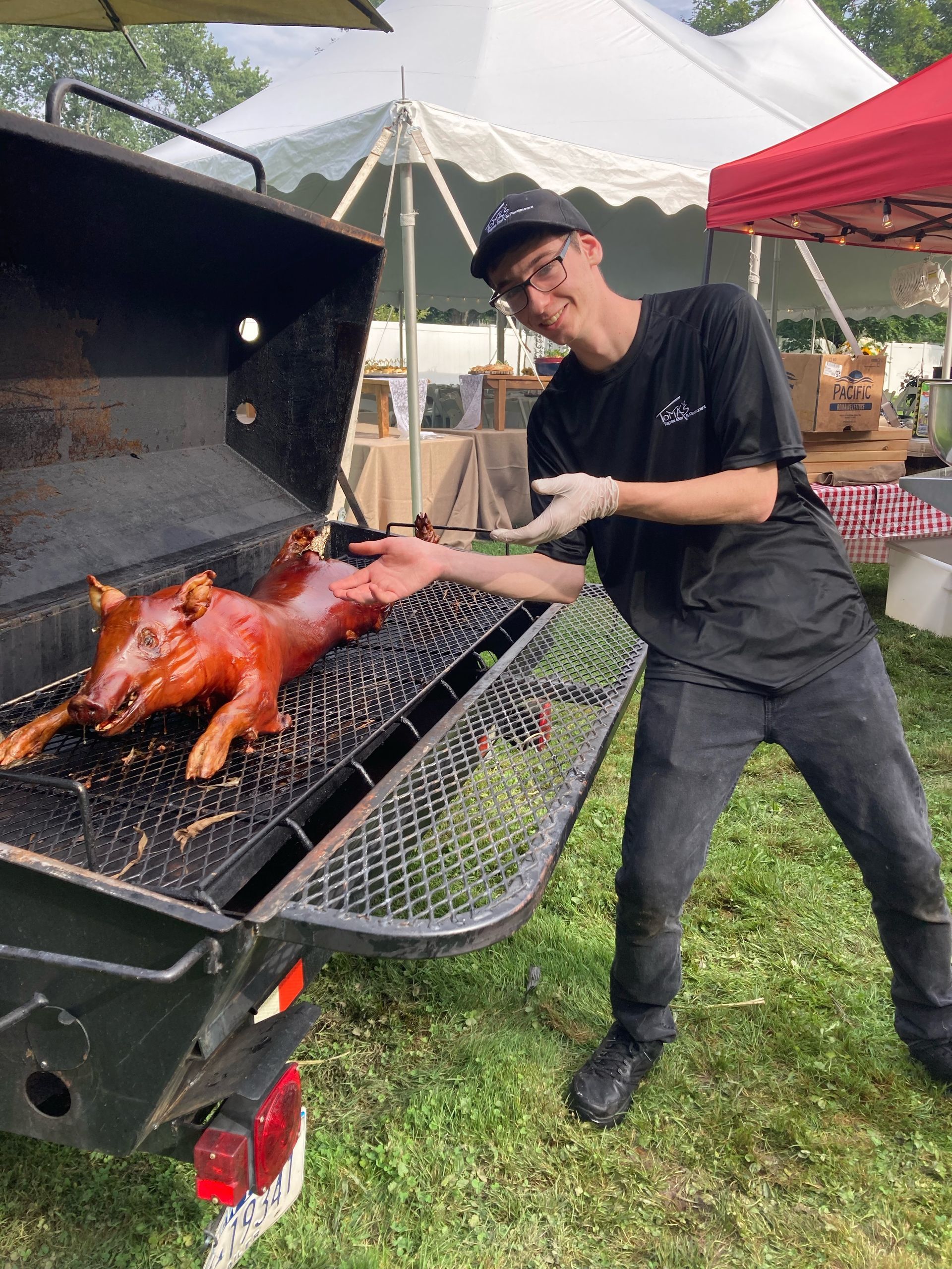 Man in black clothing gestures at roasted pig on a large barbecue grill, outdoors.