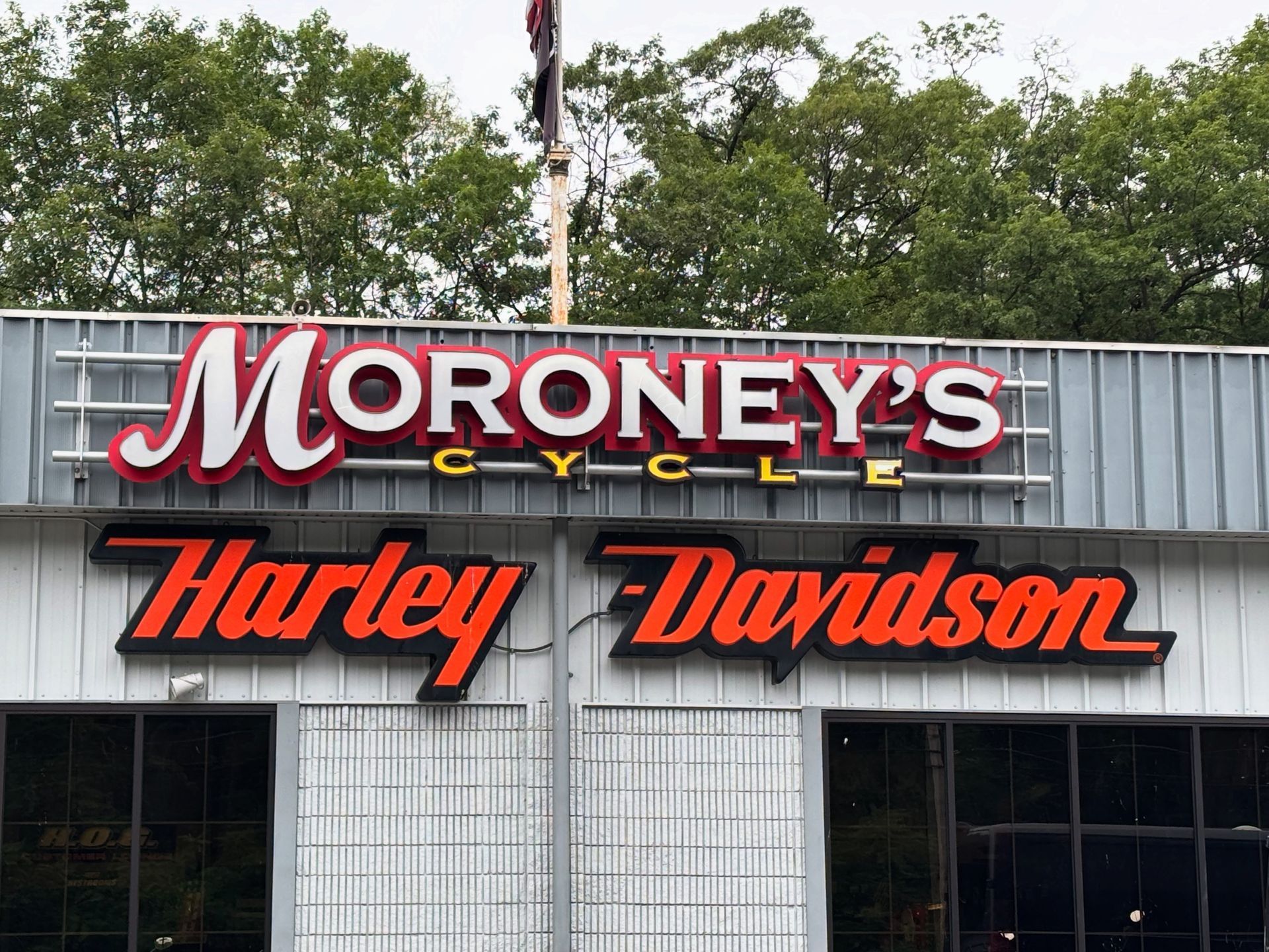 Sign for Moroney's Cycle Harley-Davidson: red and white letters, storefront, grey siding, trees in the background.