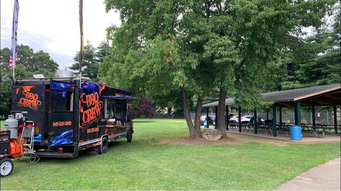 Food truck parked on grass near a picnic shelter. Trees in the background. Cloudy sky.