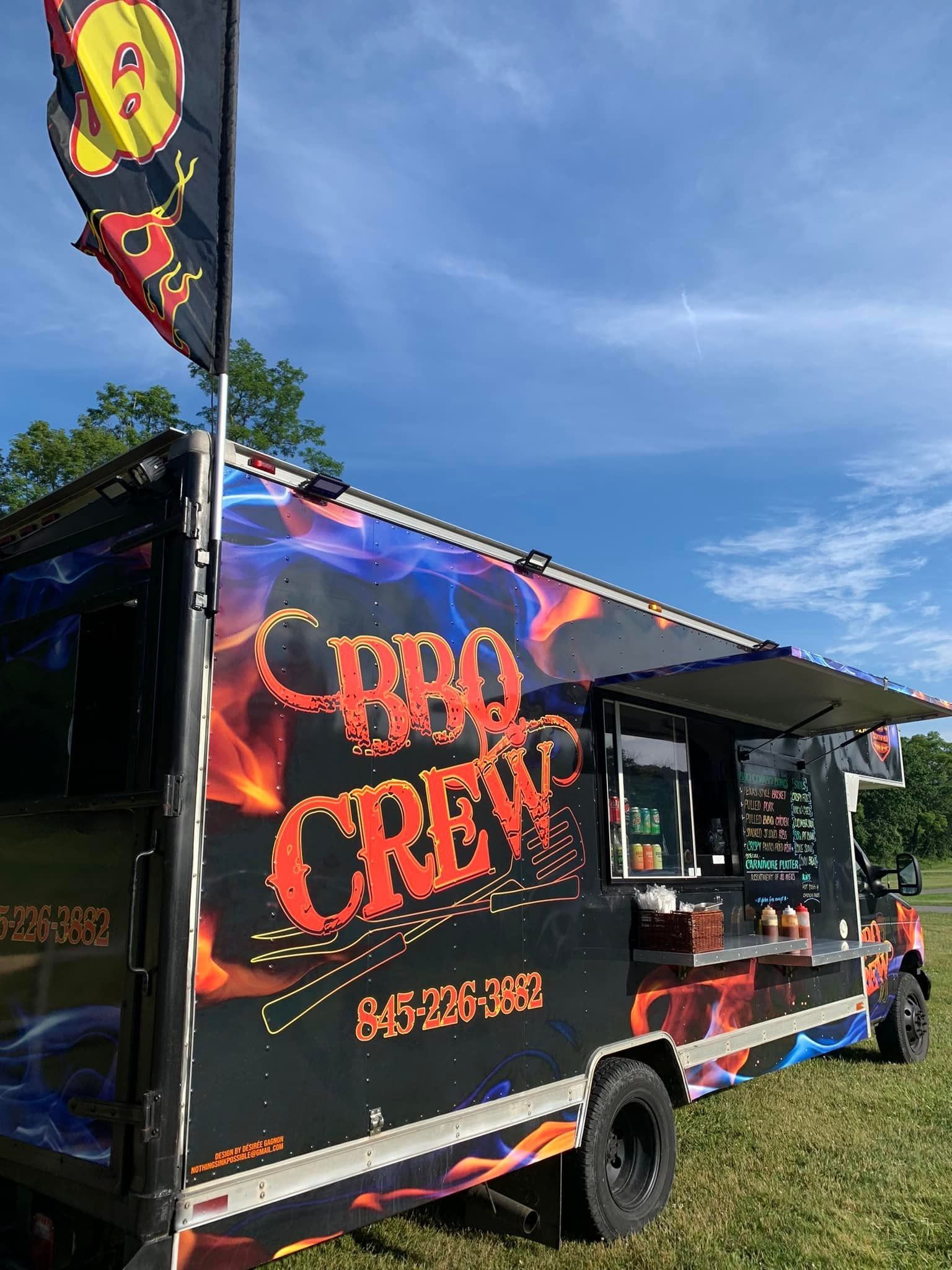BBQ Crew food truck with flame graphics, parked on grass under a blue sky, flag waving.