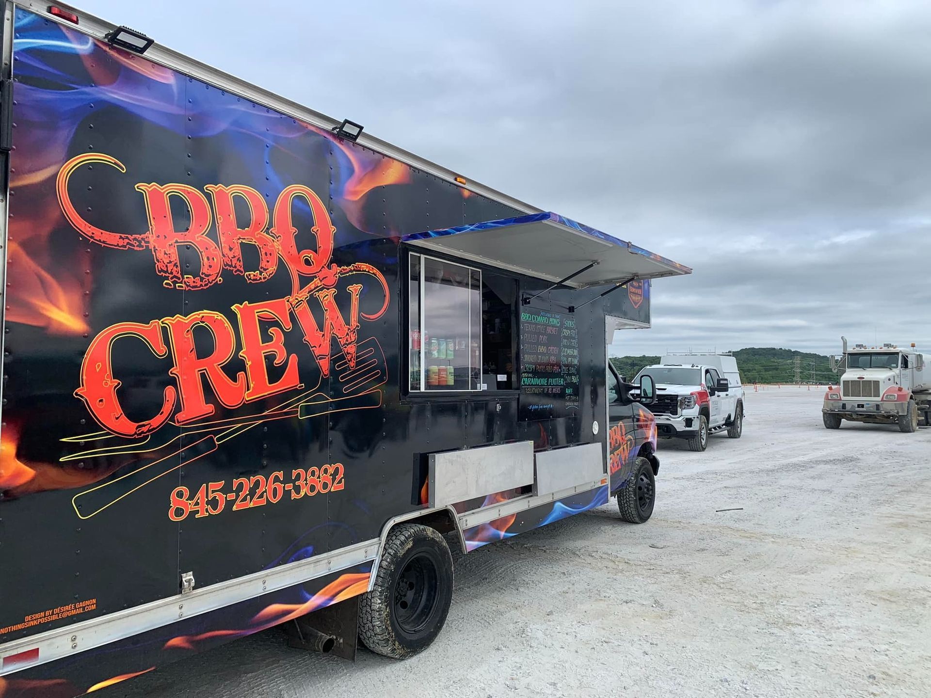 BBQ Crew food truck with flame graphics, serving window open. Trucks parked nearby under a cloudy sky.