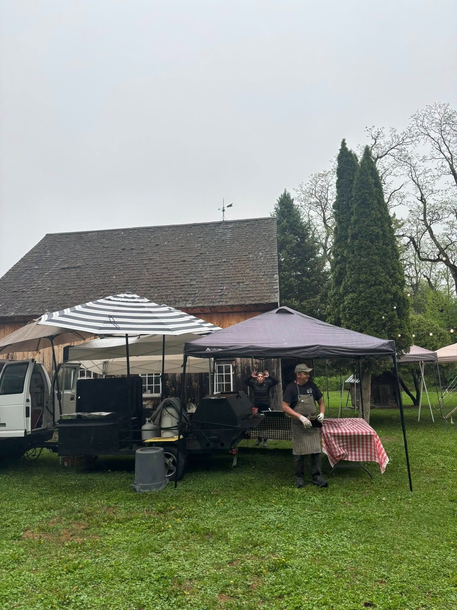 Food vendor with smoker and tents in front of a barn on a cloudy day. Man in apron.