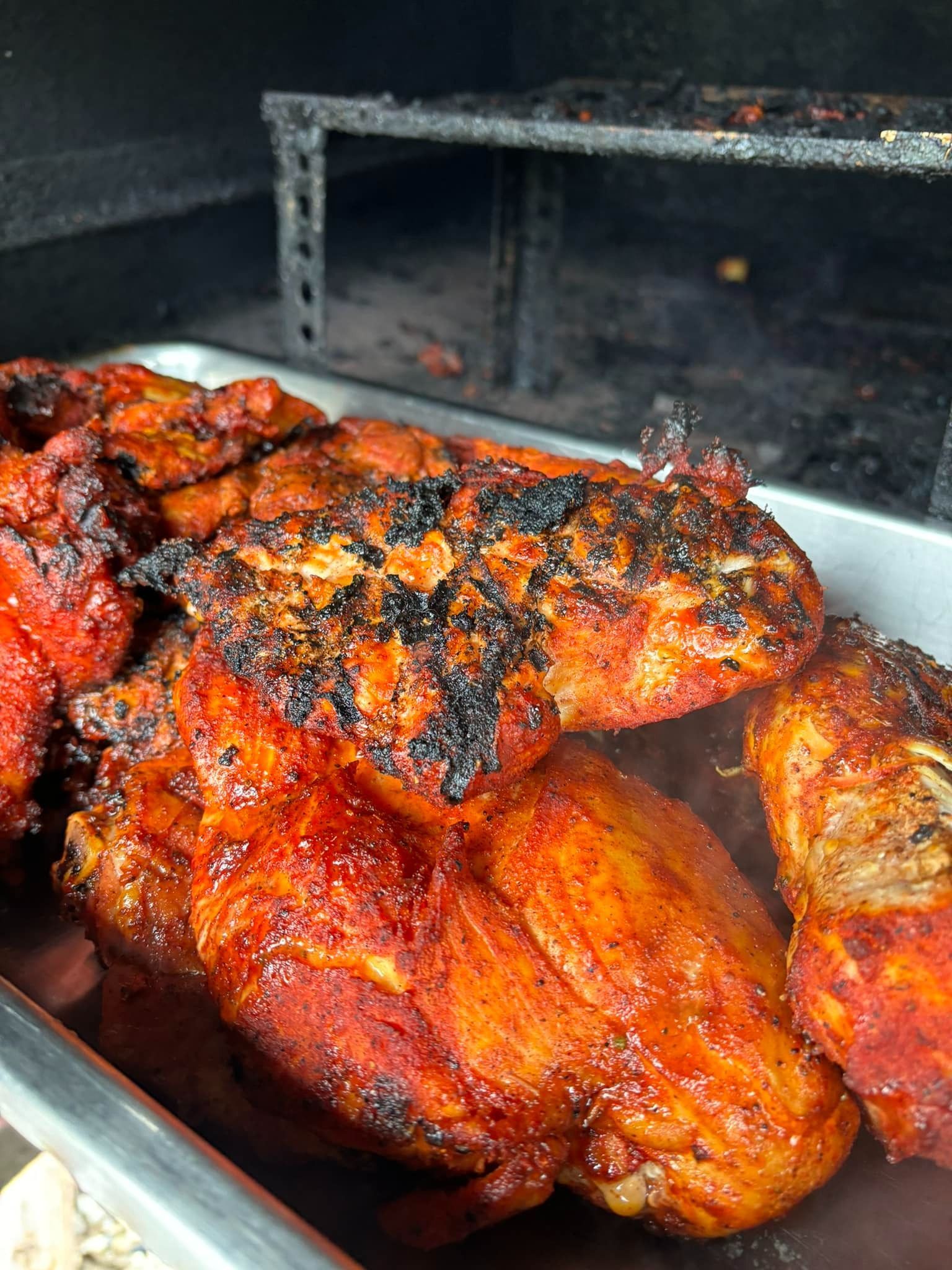 Cooked, reddish-brown chicken pieces piled in a metal tray with a smoky grill in the background.