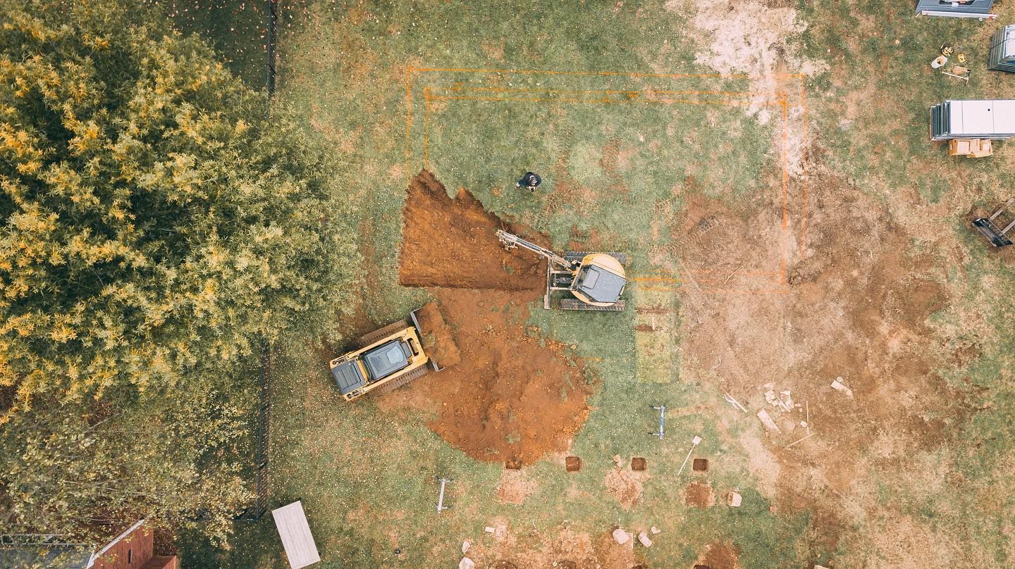 Aerial view of construction site with backhoe and excavator digging in grassy field.