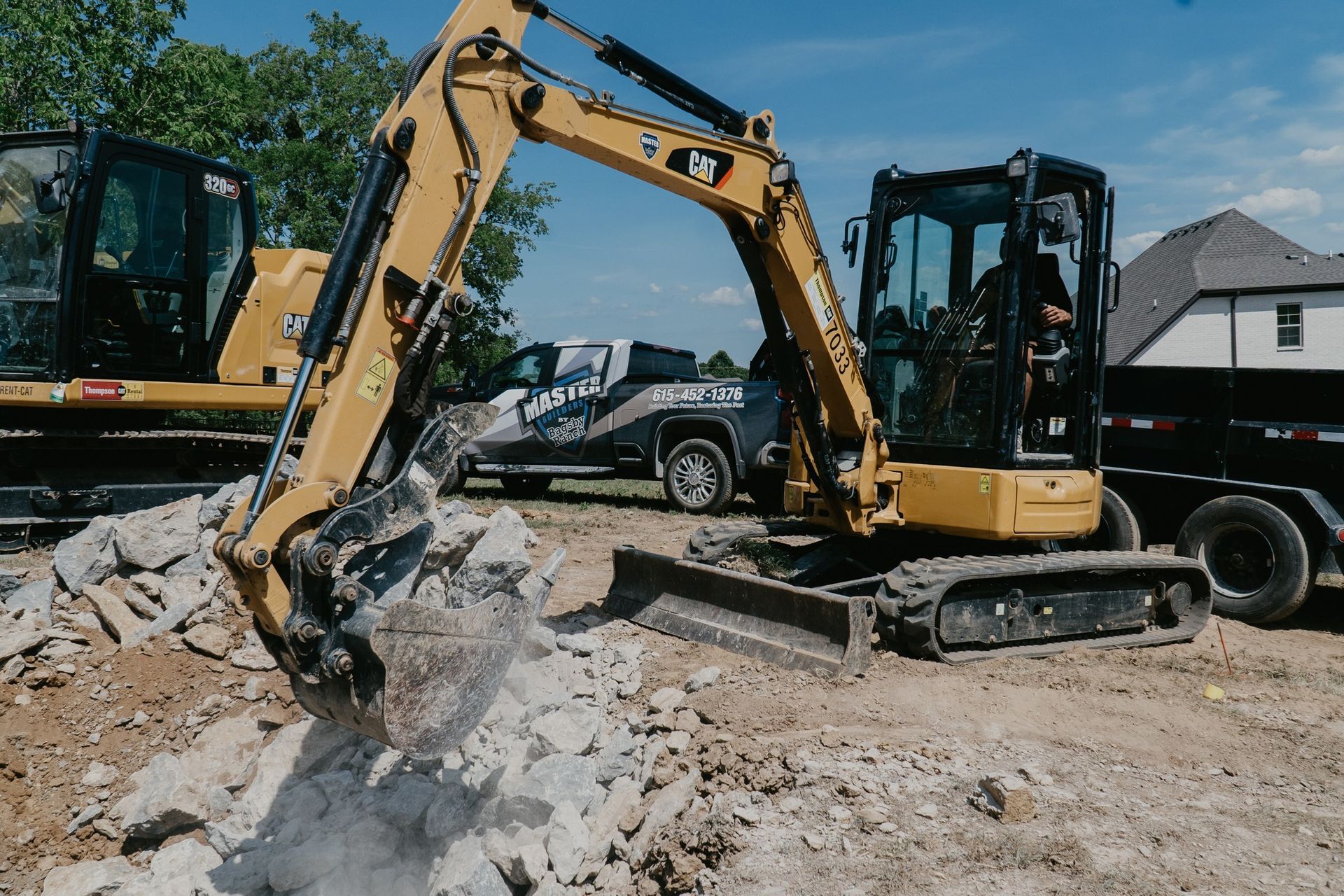 Yellow excavator digging through rubble at a construction site; a truck and trailer are in the background.