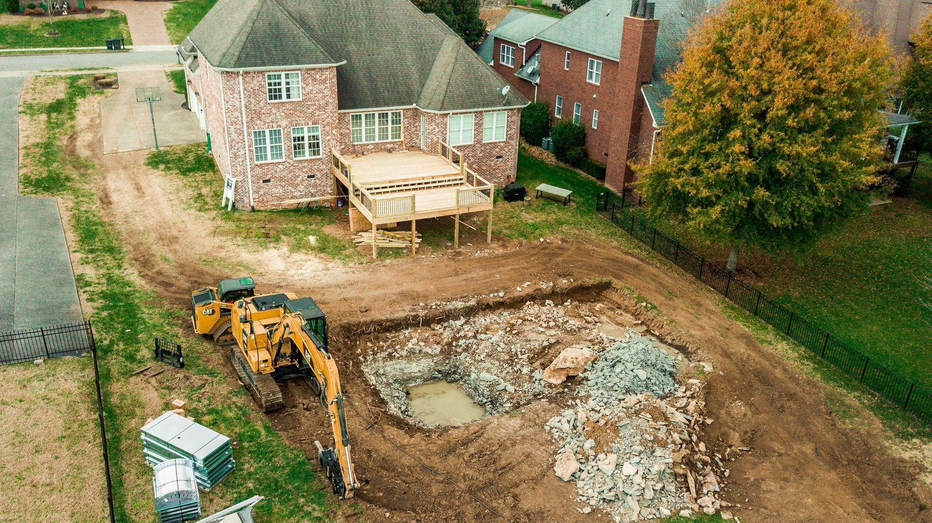Yellow excavator digs a large hole in a yard next to a two-story brick house with a wooden deck; a construction site.