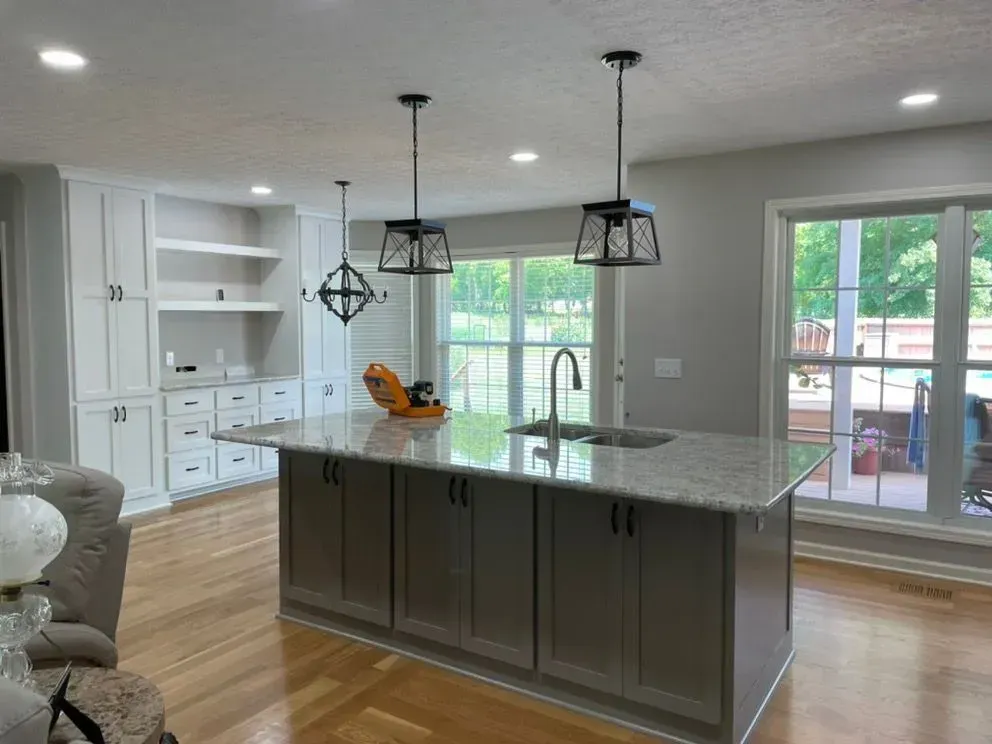 Kitchen with gray island, white cabinets, and pendant lights; view of a yard through a large window.