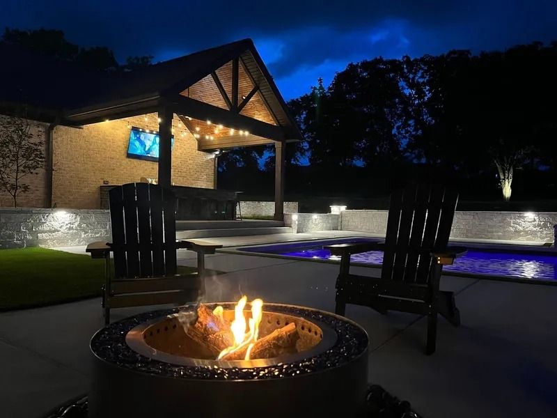 Night scene: patio with lit fire pit, chairs, pool, and covered outdoor space.