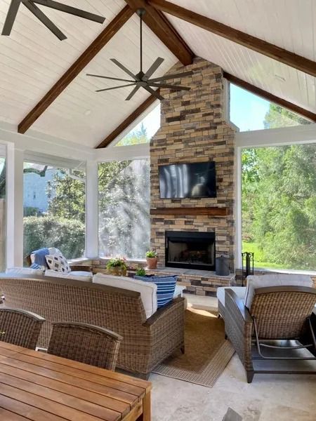 Sunroom with stone fireplace, glass windows, exposed beams, wicker furniture, and a ceiling fan.