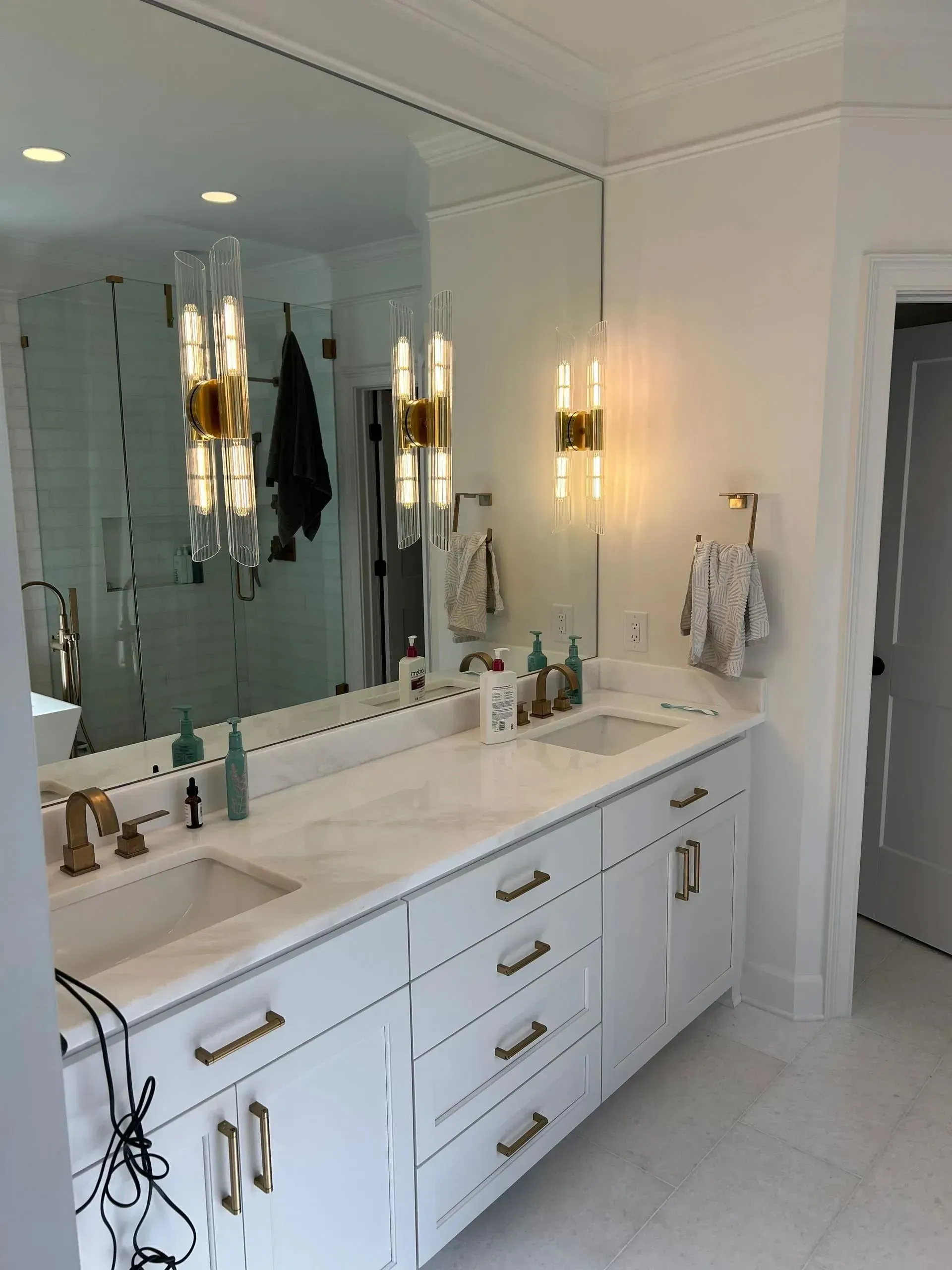 White bathroom with marble countertop and large mirror; two sinks with gold fixtures, two sconces.