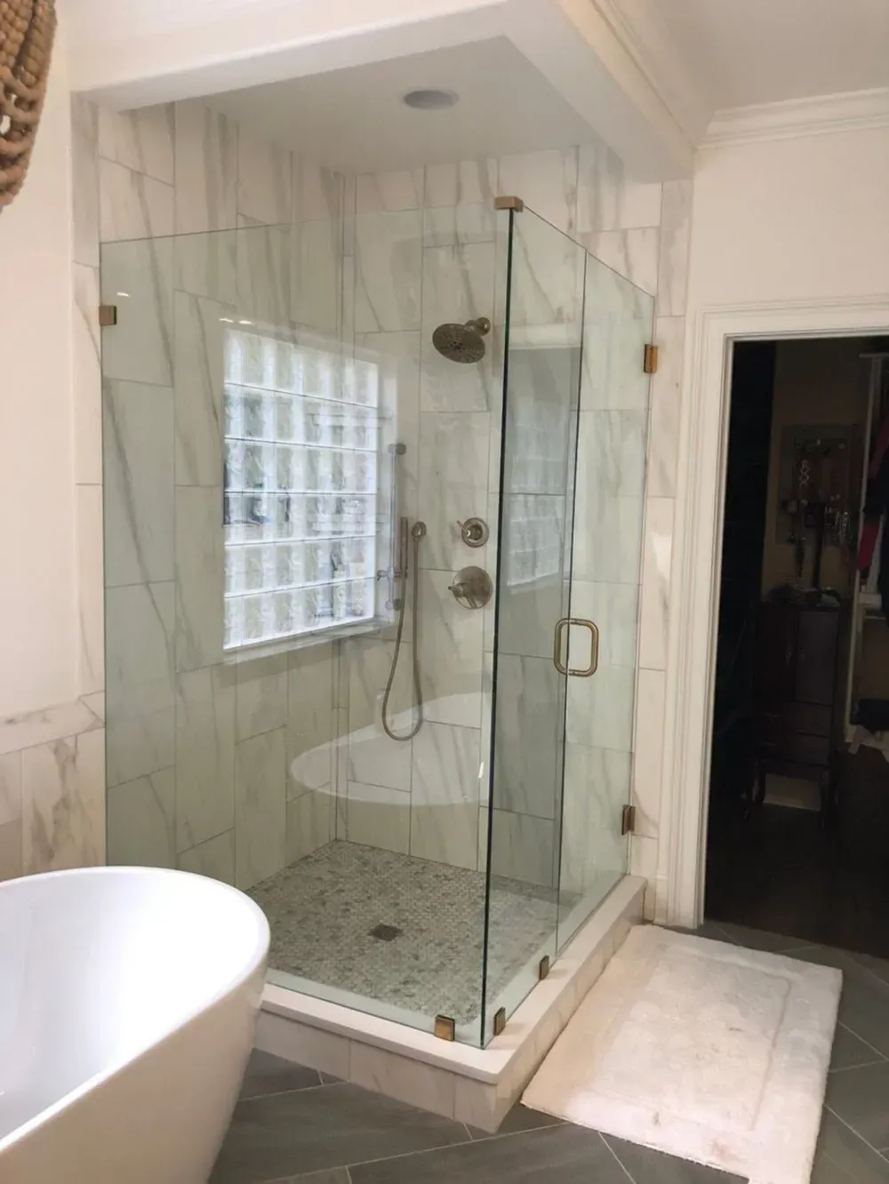Glass shower enclosure in a bathroom with white marble tile, gold fixtures, and a glass block window.