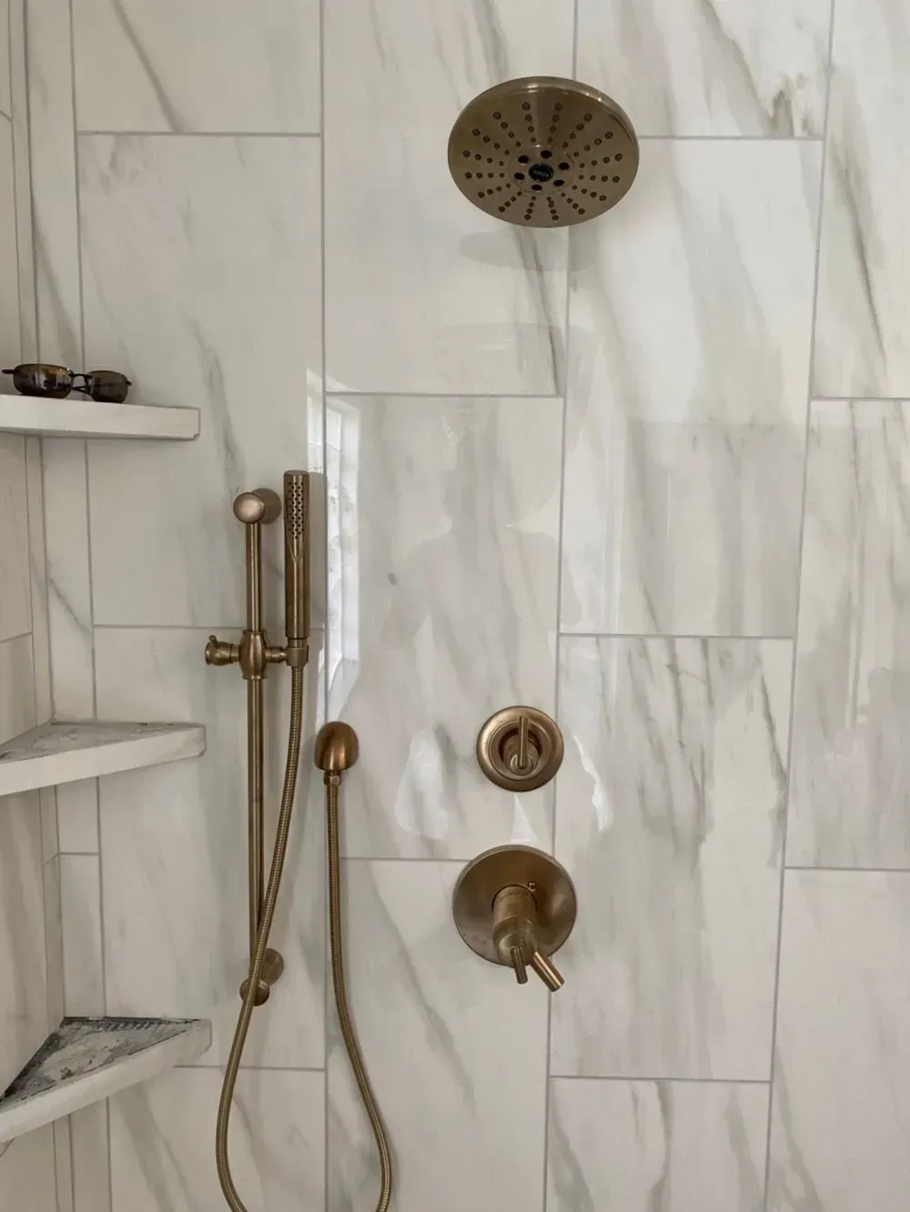 Shower with marble-look tile, featuring bronze fixtures: showerhead, handheld, and water controls. Built-in shelves.