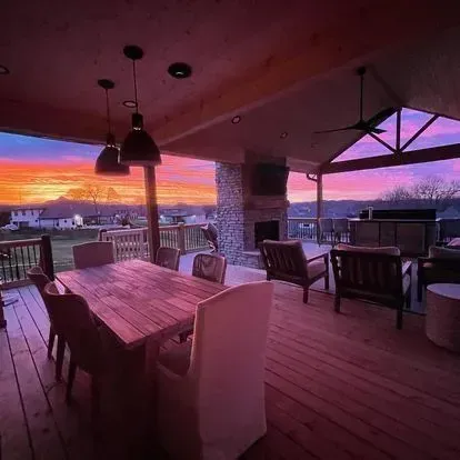 Outdoor patio with a wooden deck, dining table, and fireplace, under a sunset sky.