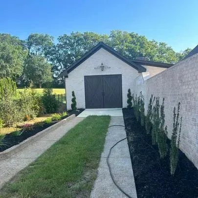 White stucco garage with brown doors, black roof, and adjacent brick wall. Lawn and shrubs in front.
