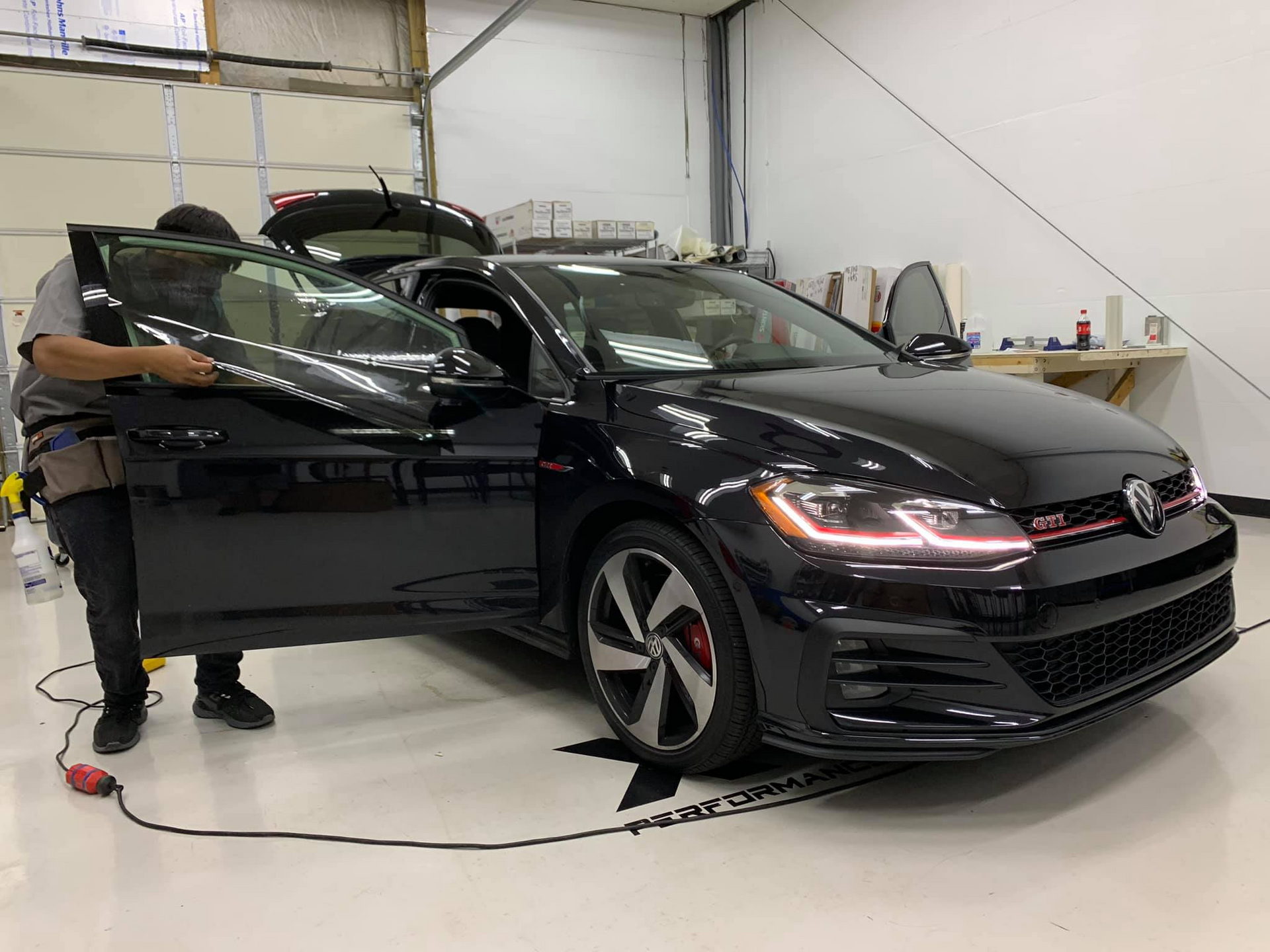 A man is applying window tinting to a black car in a garage.