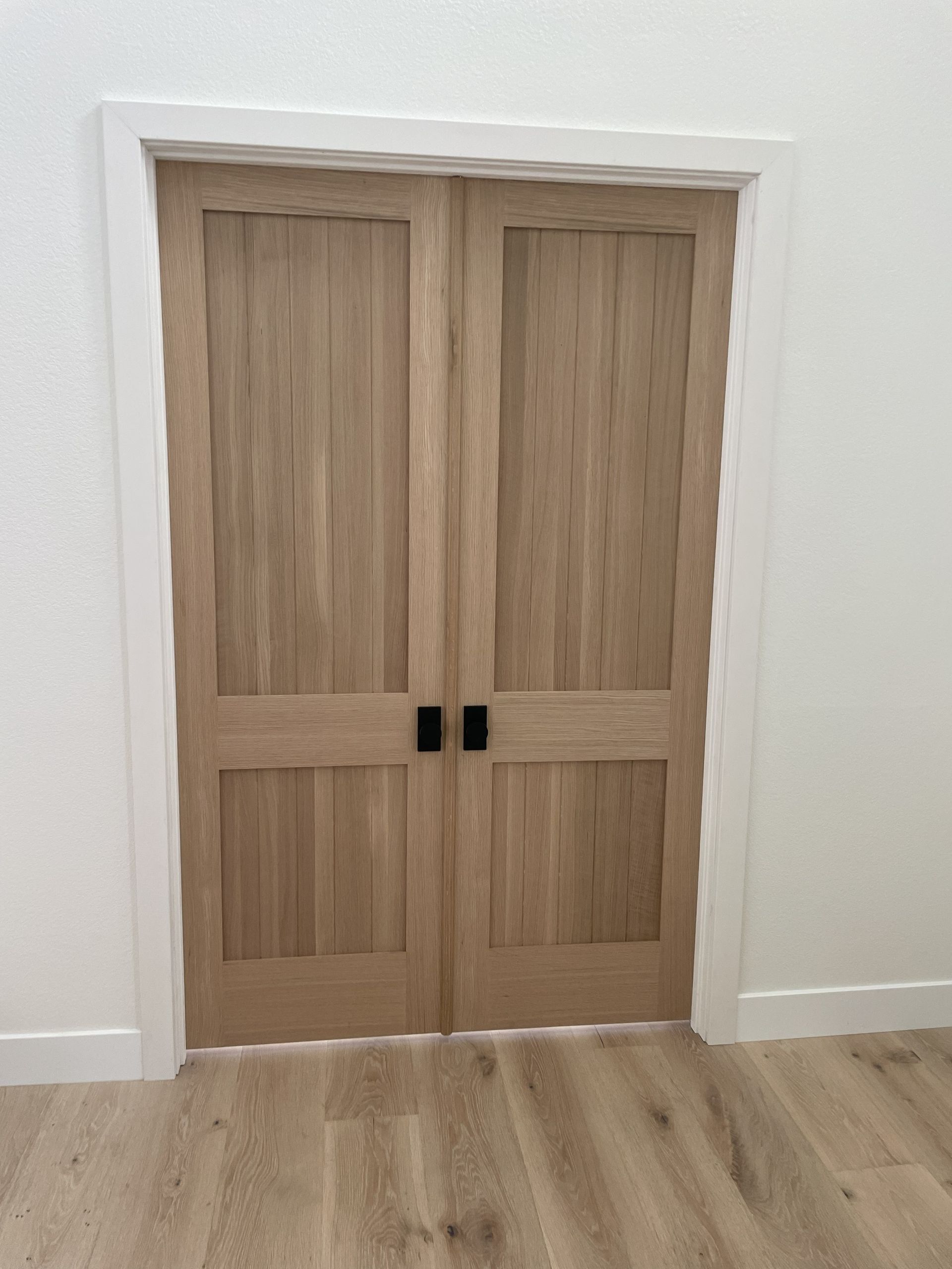 Double wooden doors with black handles in a white hallway with light wood flooring