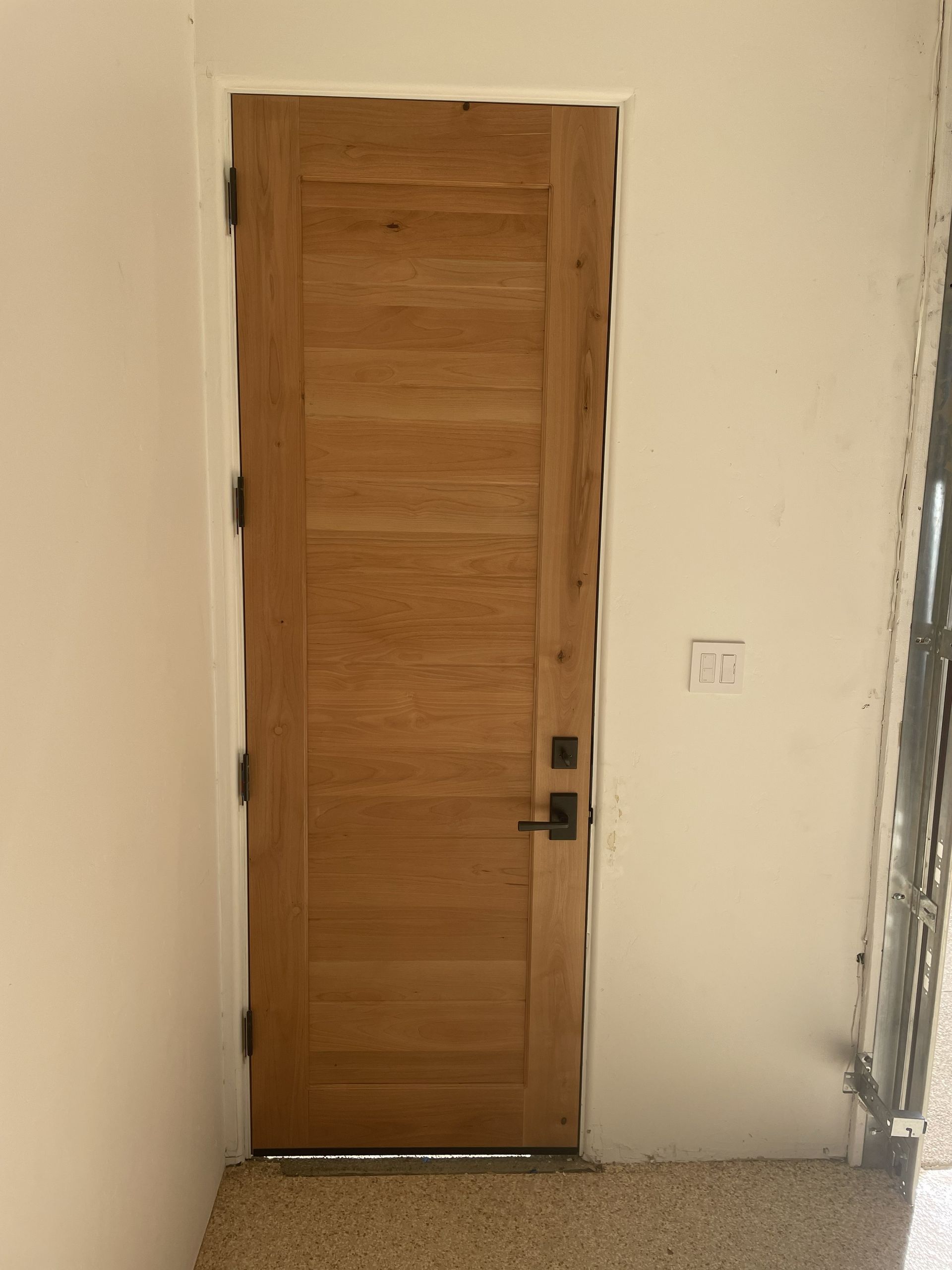 Wooden front door in a narrow hallway with white walls and a tiled floor