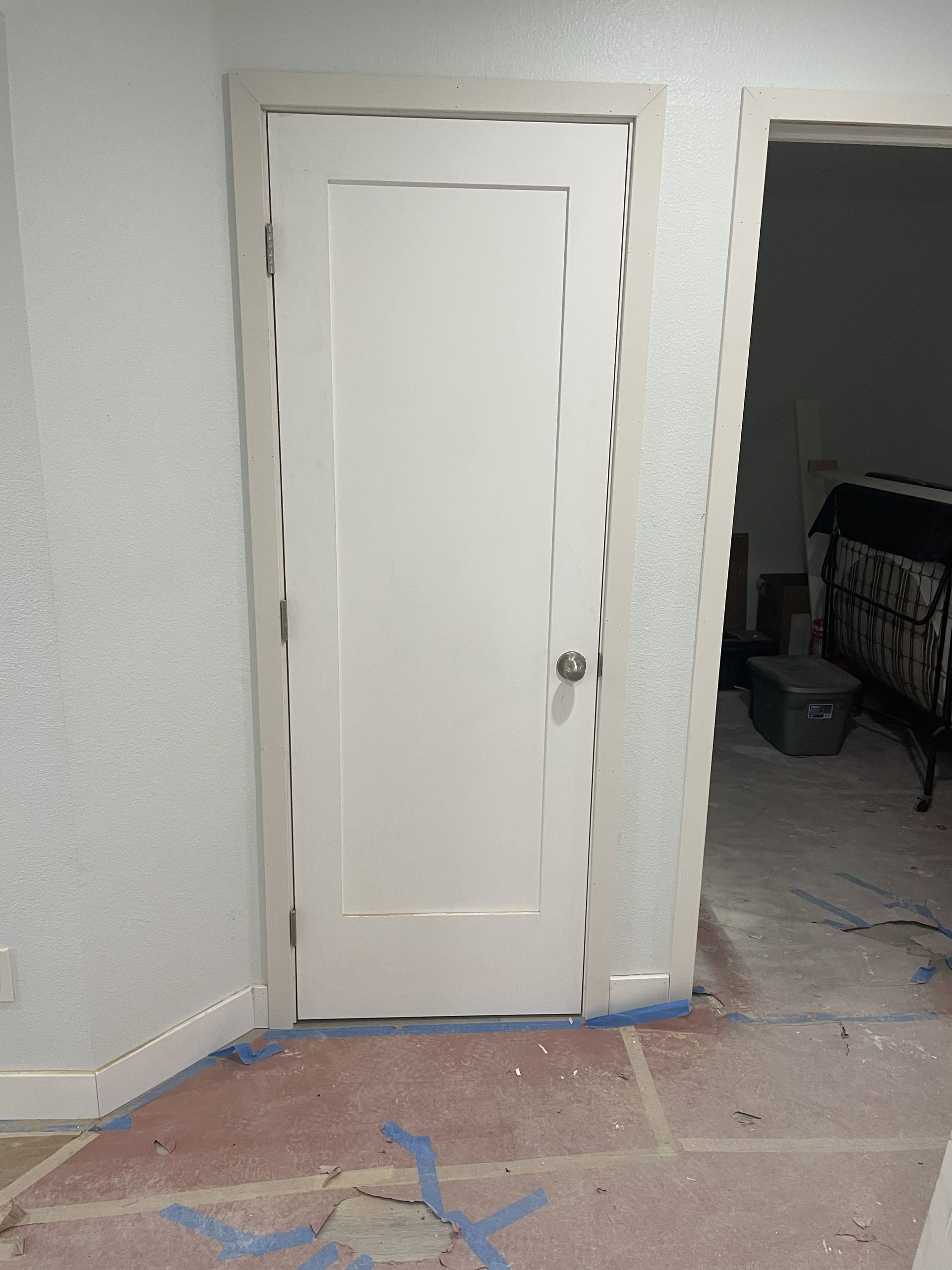 White interior door in a room under renovation, with taped floor and open doorway to another room.