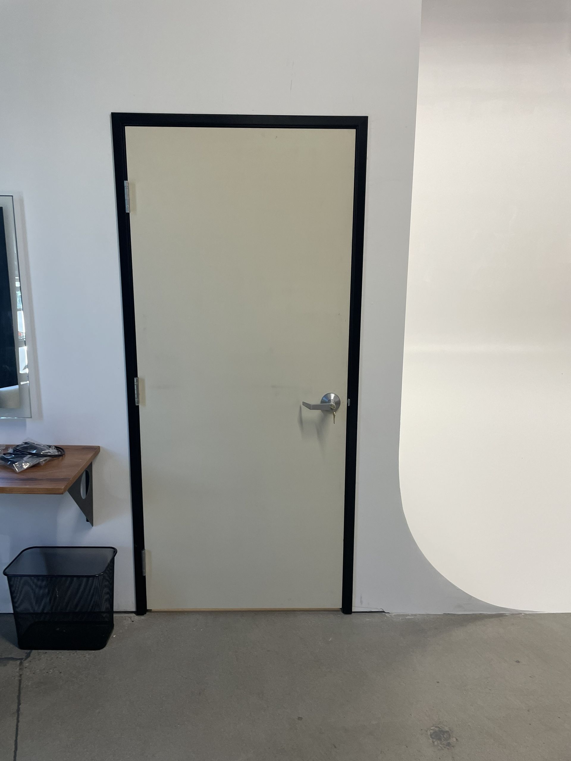 Closed white door with black trim on a gray wall, next to a small table and black trash bin.