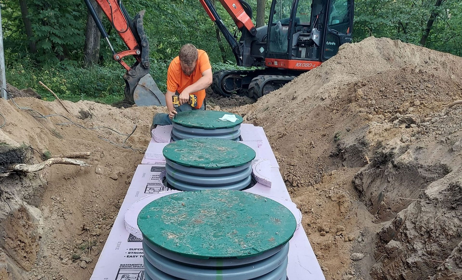 Man installing septic tank lids in a trench with an excavator in the background.