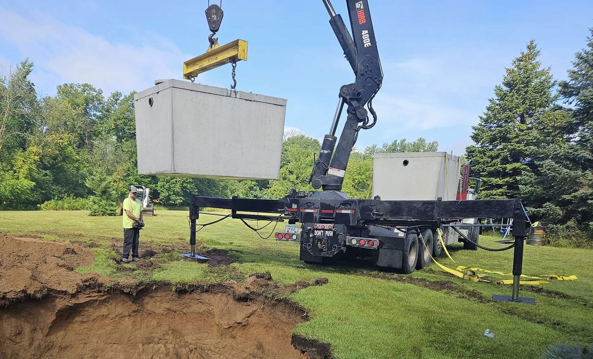Crane lifting a concrete septic tank over a large excavated hole on a grassy lawn.
