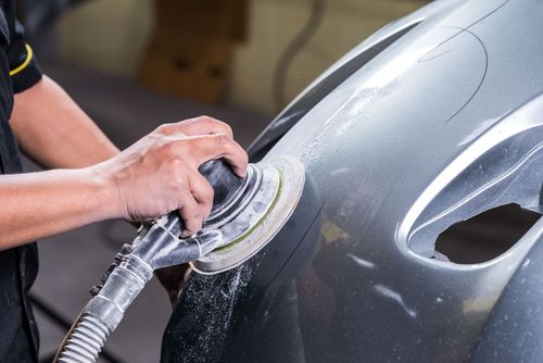 Hands sanding a gray car body with a power sander in an auto shop