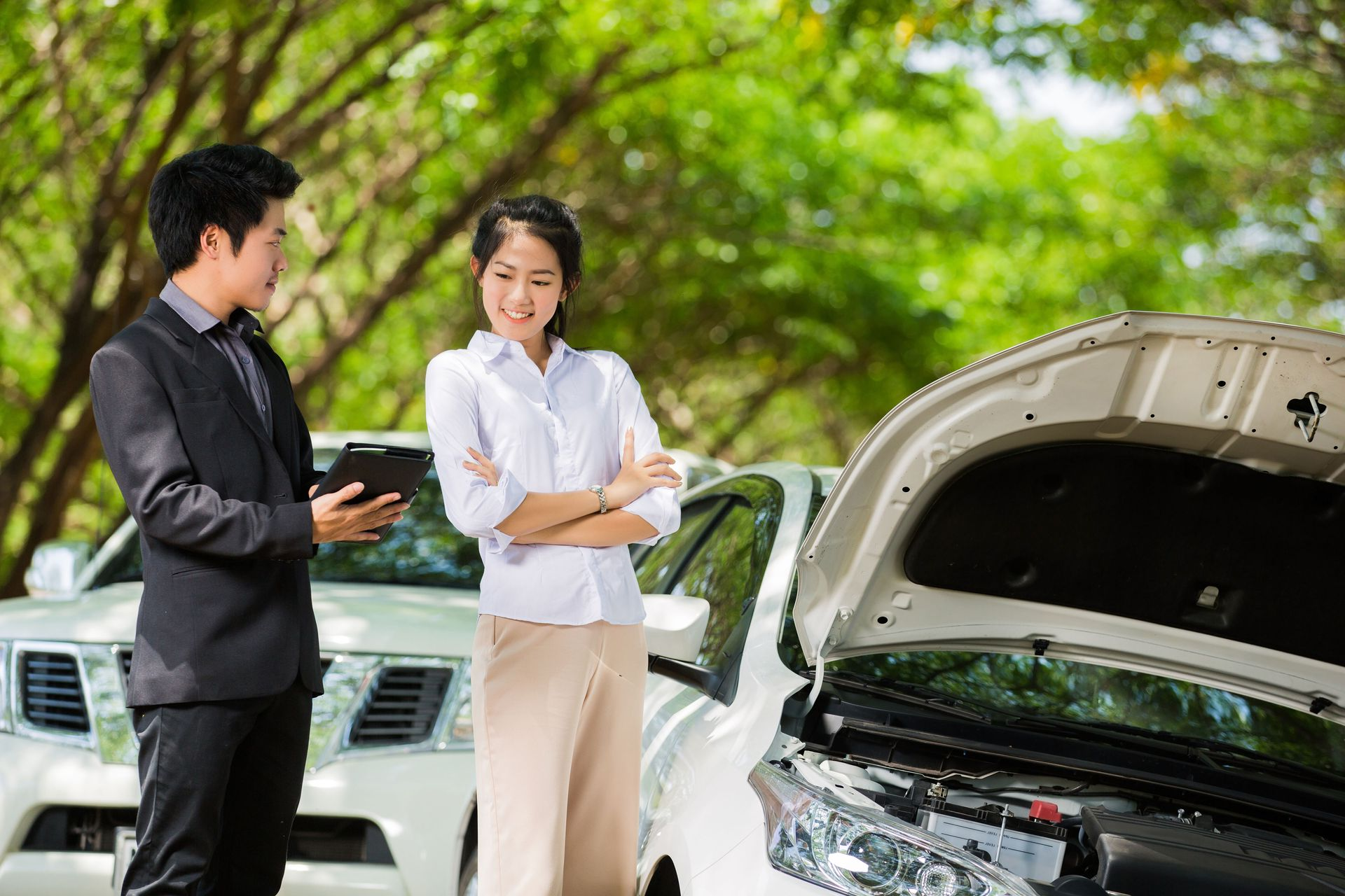 Two people discussing a white car with its hood open in a green outdoor setting