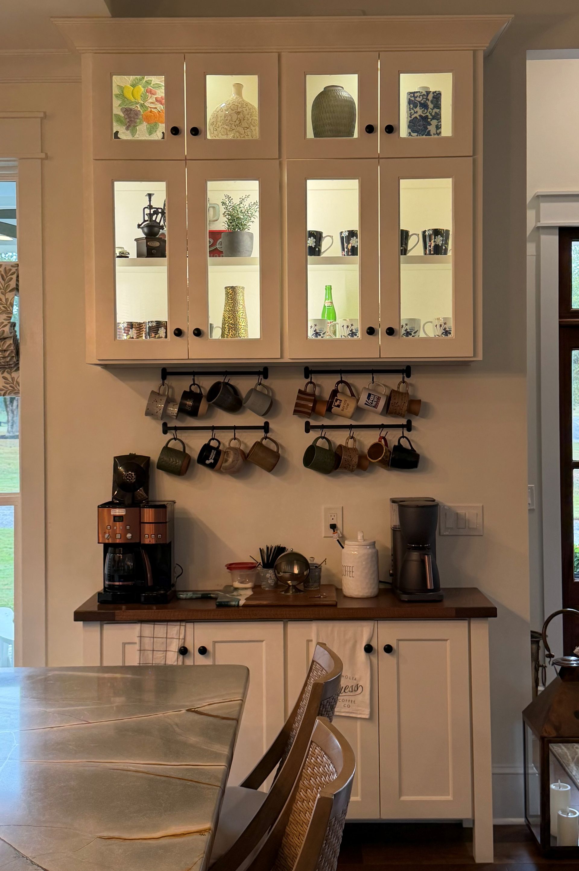 Kitchen with white cabinets, black countertop, hanging mugs, and a coffee maker near a window.