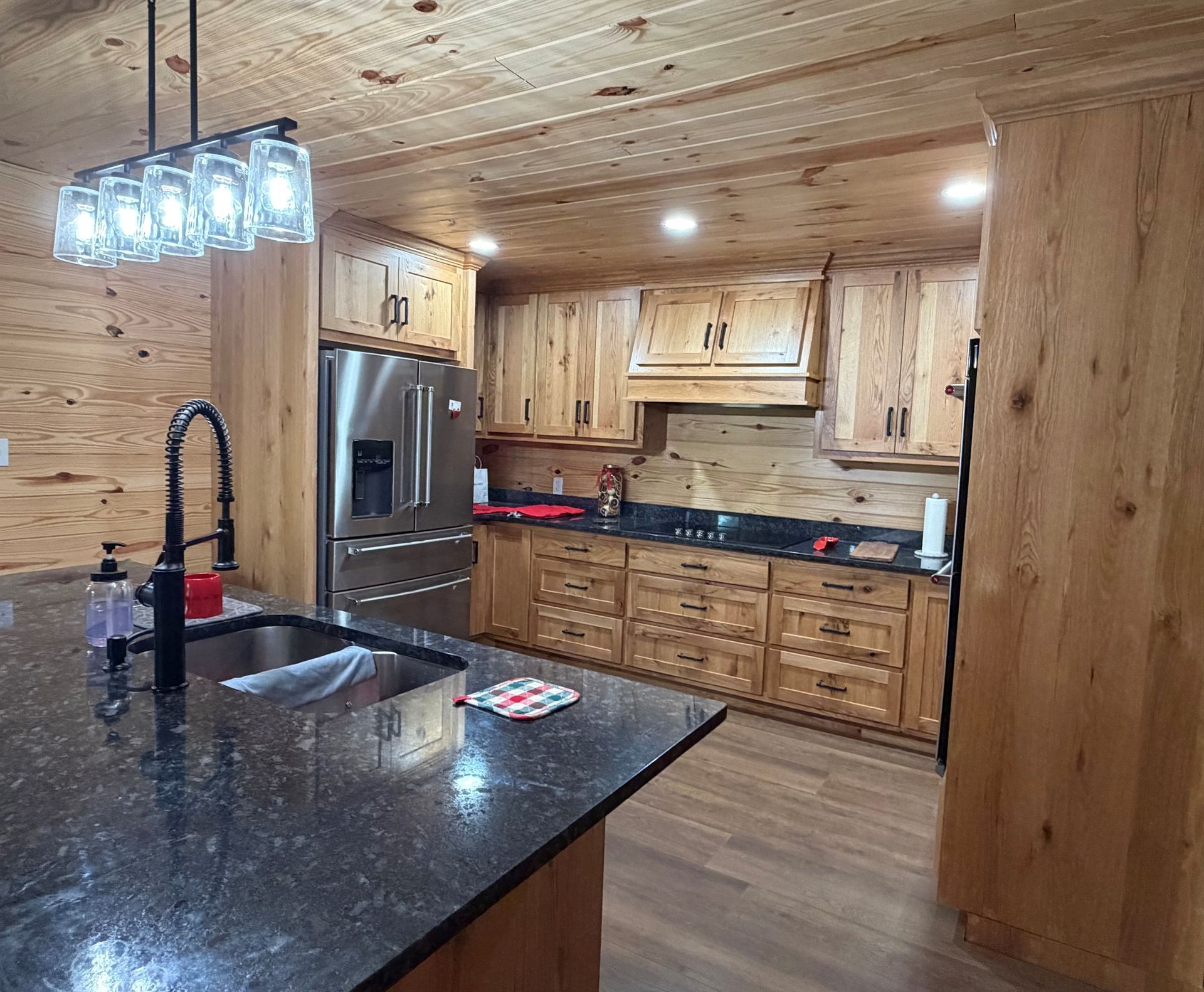 Rustic kitchen with wood cabinets, black countertops, stainless fridge, and island pendant lights