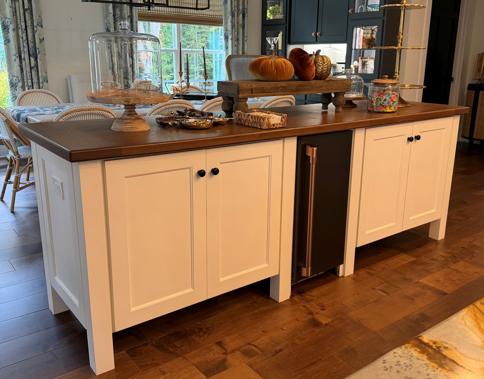 White kitchen island with wood countertop, cabinets, open wine fridge, and decorative bowls in a bright dining area