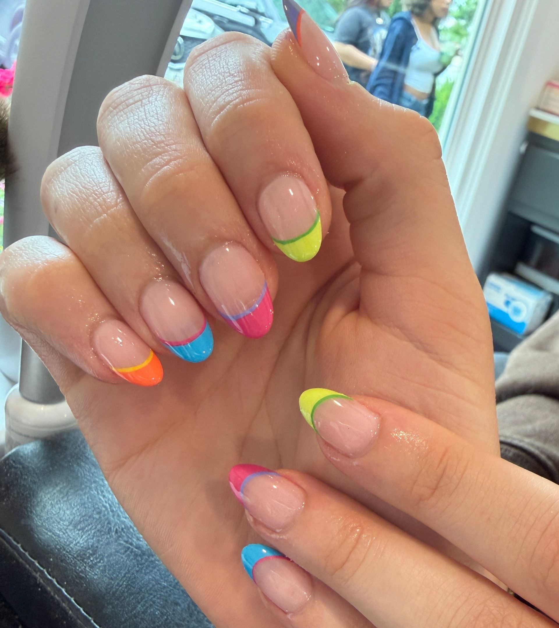 Hand with glossy French-tip nails in neon pink, blue, orange, and lime green.