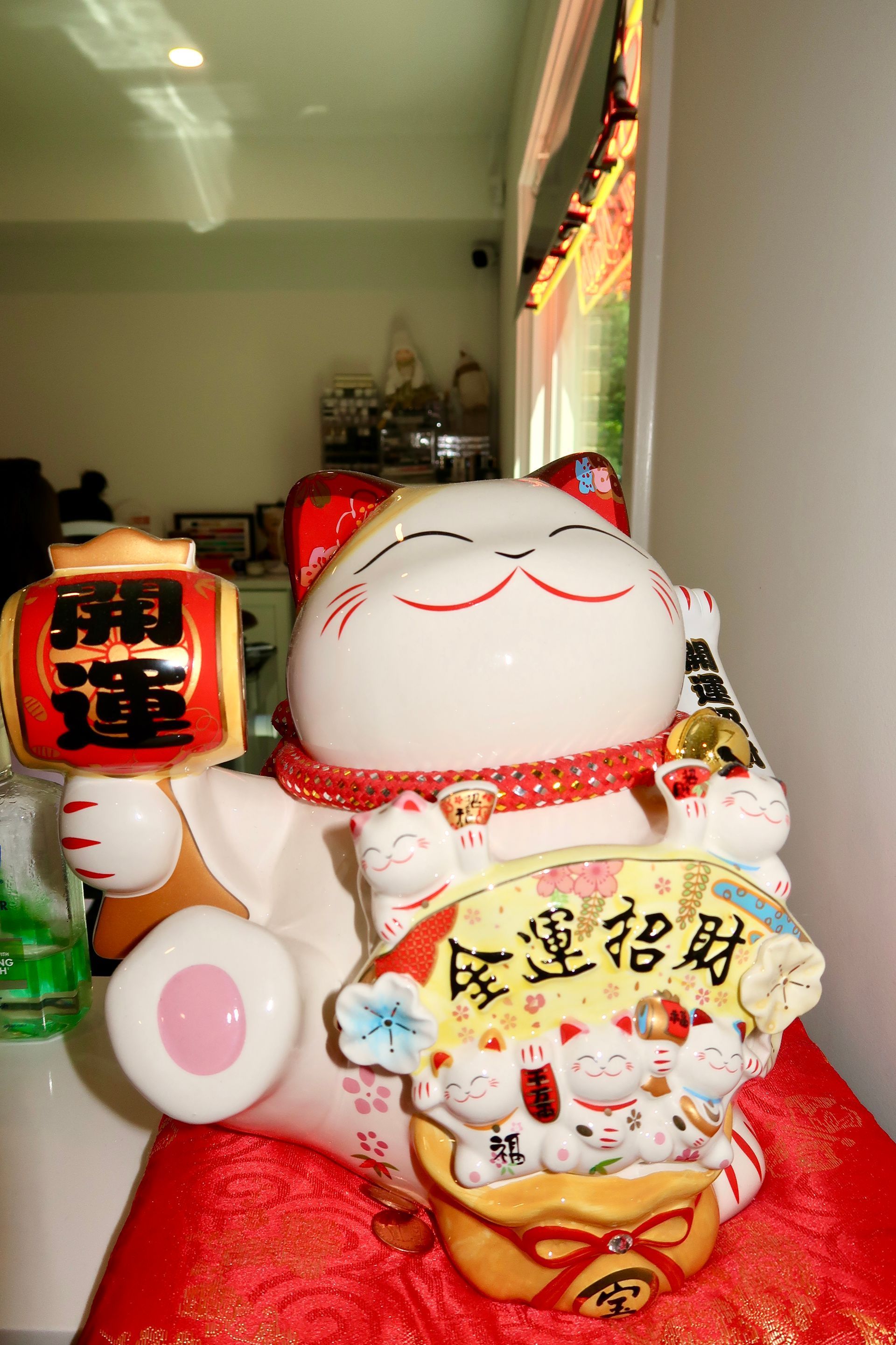 Three smiling lucky cat figurines on a red cloth by a window, including one with Chinese characters.