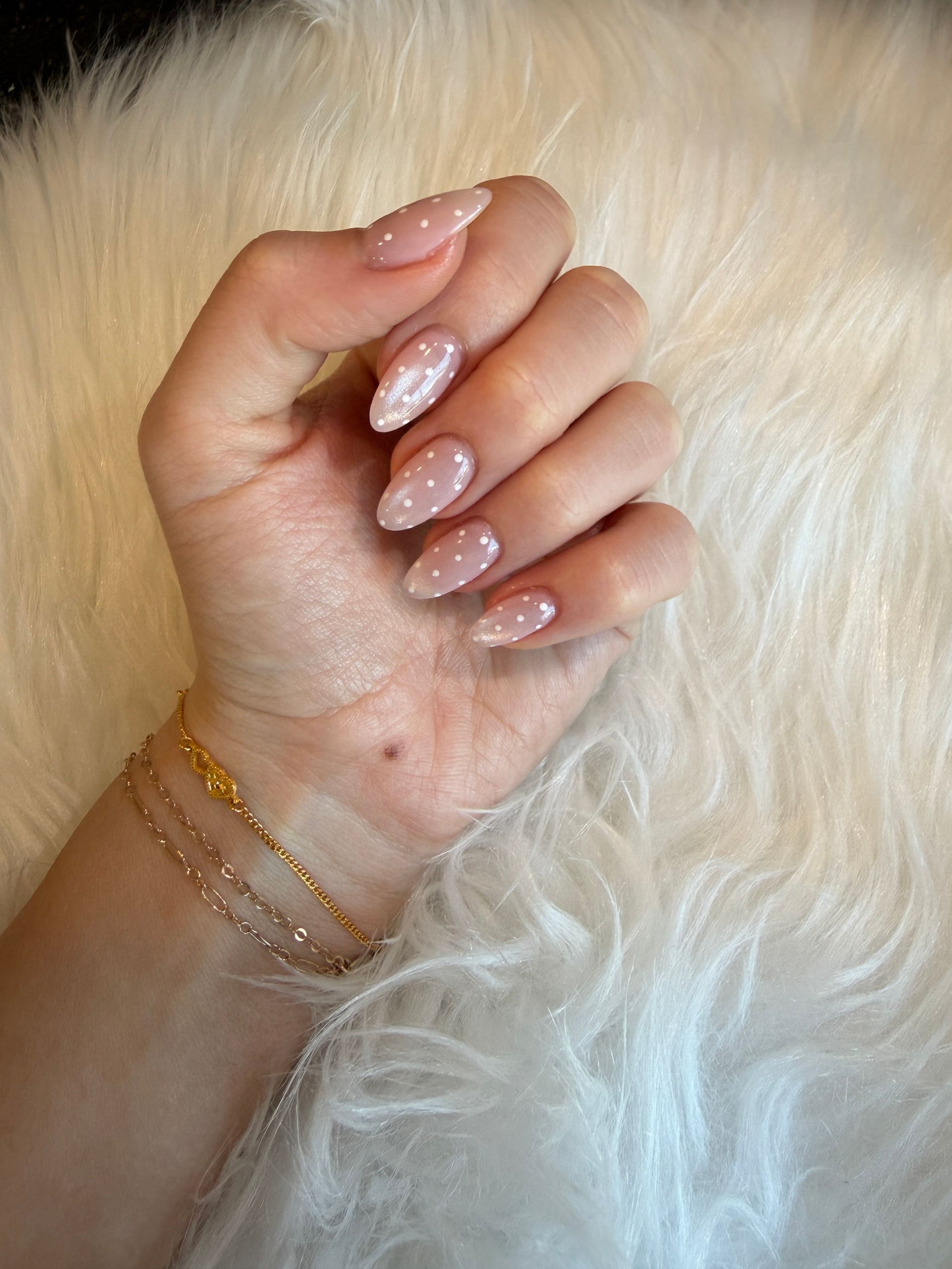 Hand with nude glitter manicure resting on white fluffy fabric, wearing delicate gold bracelets