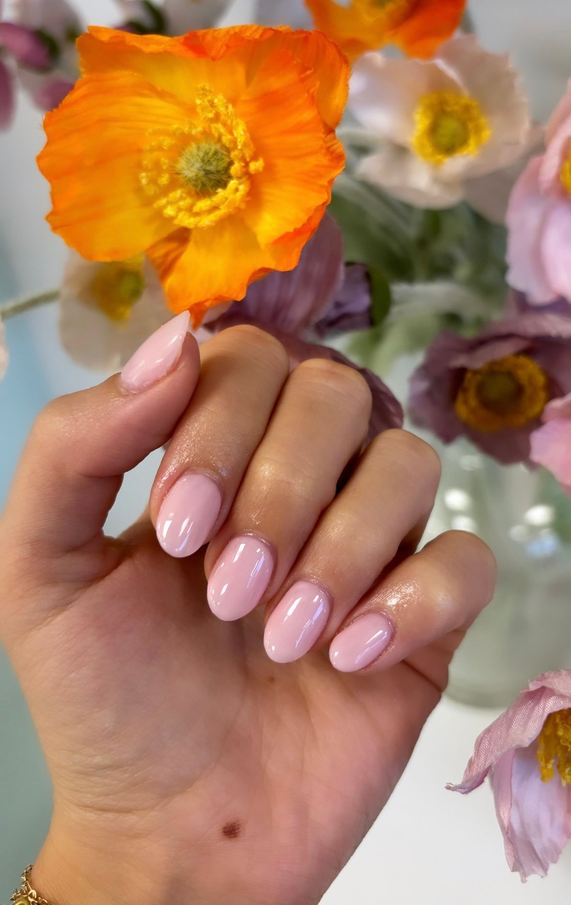 Hand with glossy pale pink nails holding orange and white flowers against a soft floral background