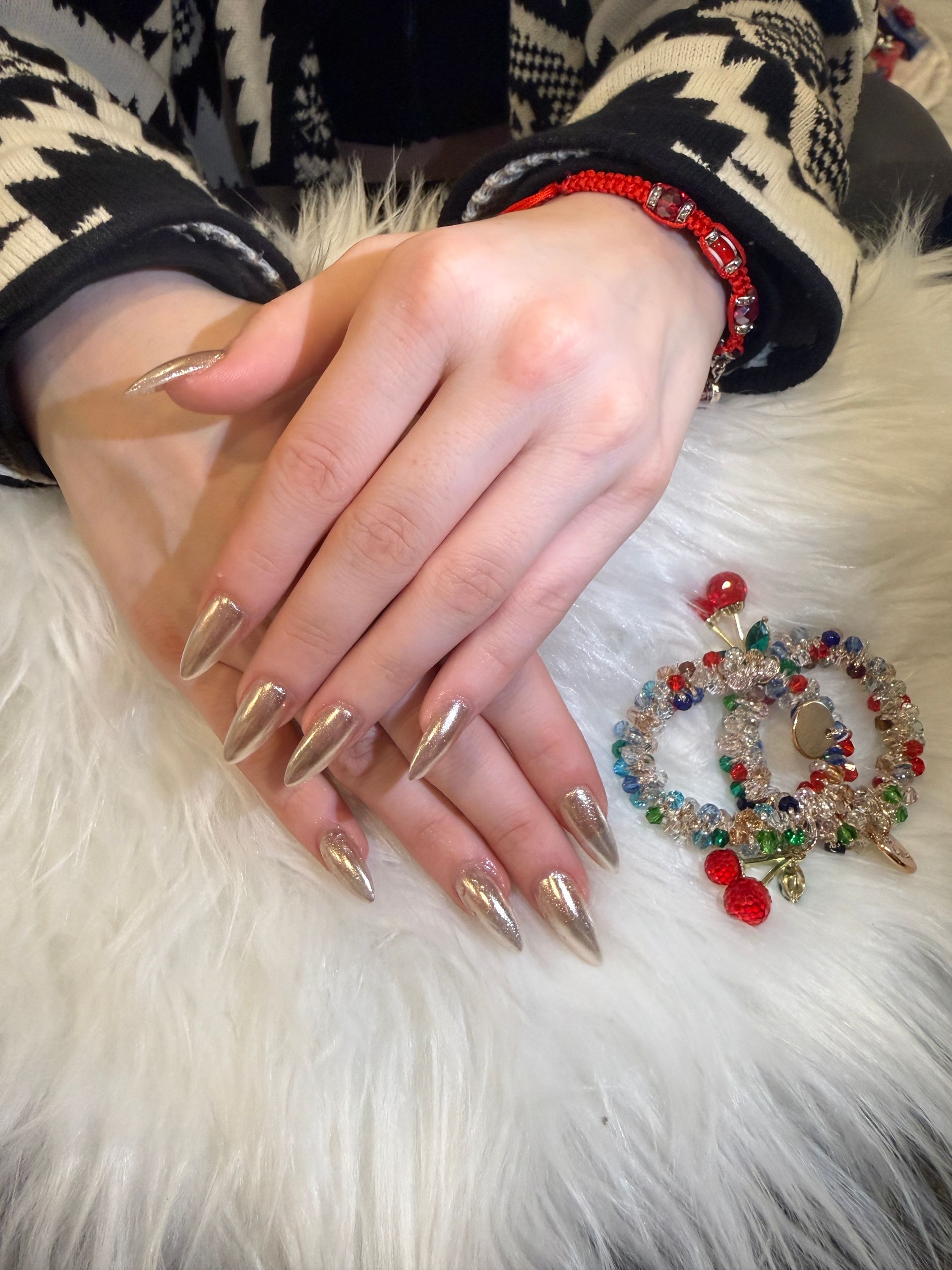 Hands with long gold nails and a red bracelet resting beside a beaded Christmas ornament on a white fur blanket