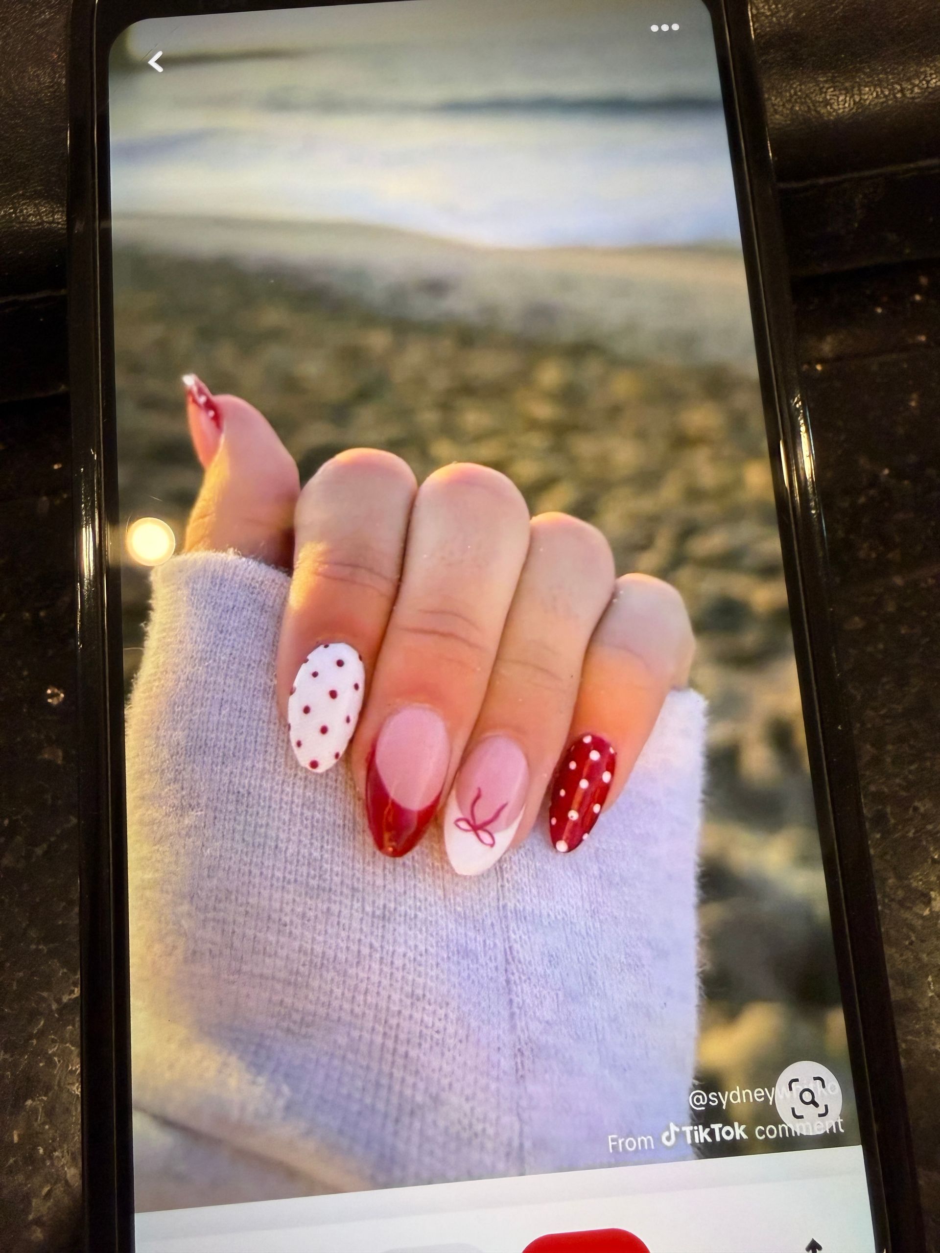 Hand with long red and white manicured nails holding a sweater sleeve outdoors on a beach.