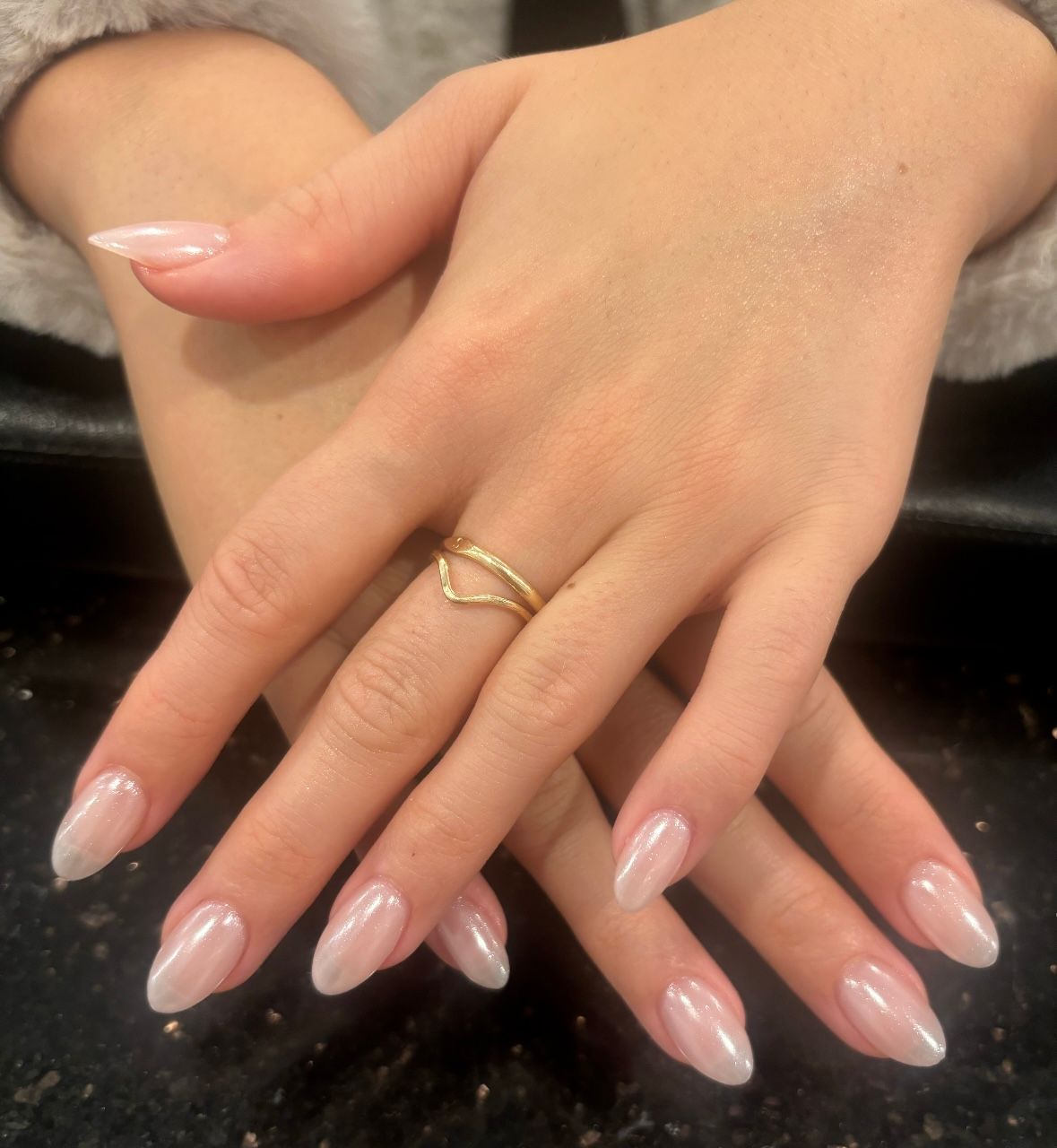 Hands with long nude-pink manicured nails and a gold ring resting on a dark surface