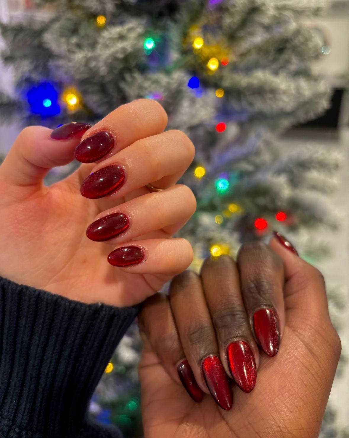 Two hands with glossy red manicures, one deep burgundy and one metallic red, in front of a lit Christmas tree