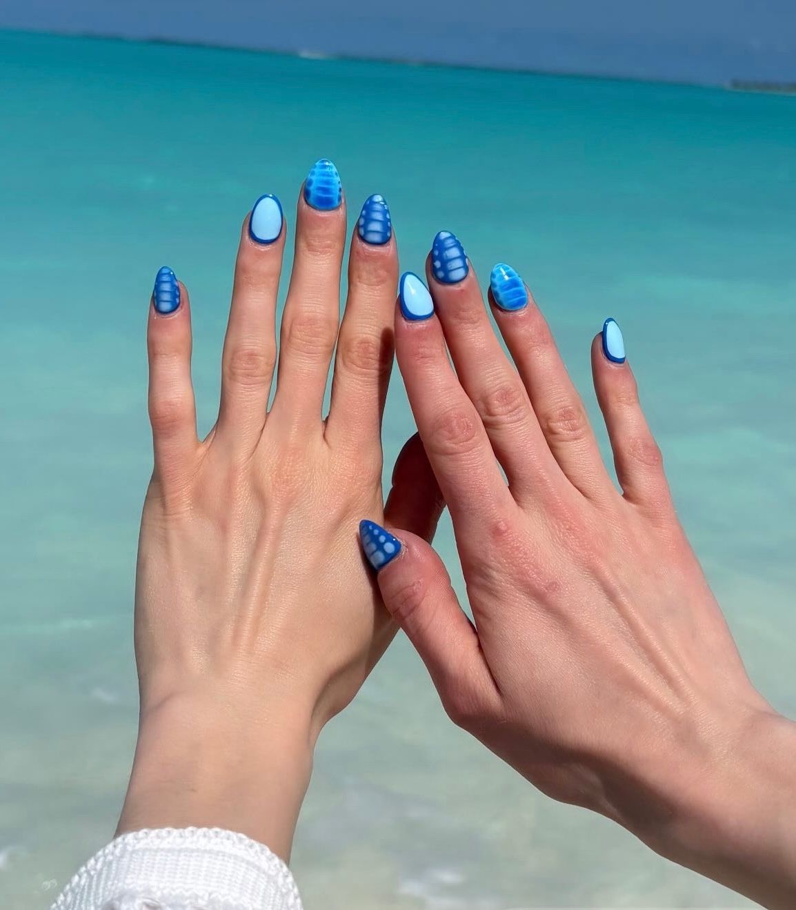 Two hands with blue manicured nails raised against a turquoise beach and ocean background