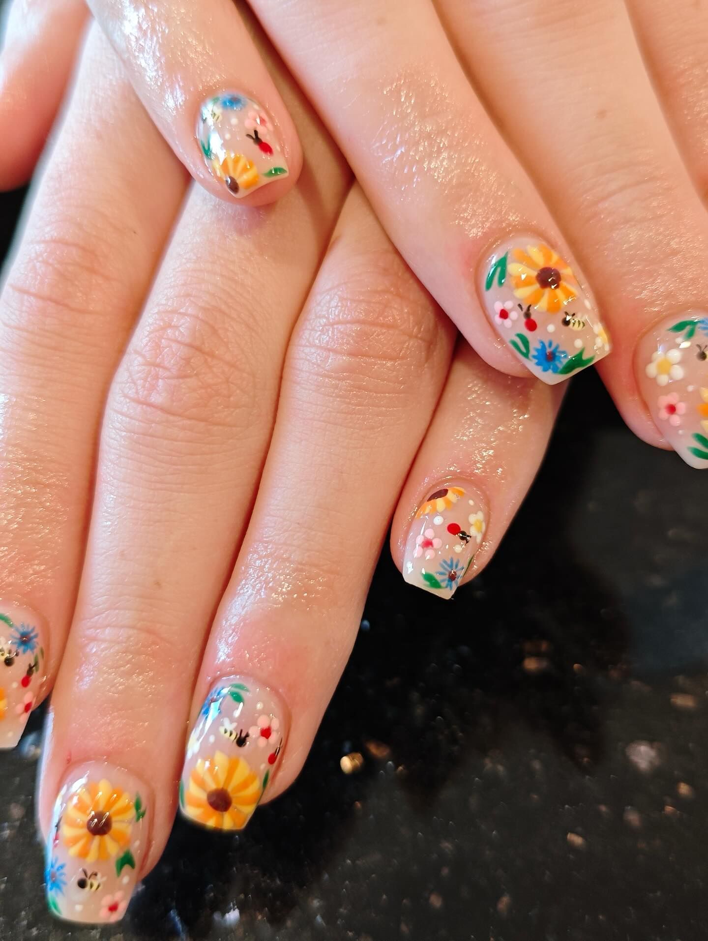 Hands with clear, shiny nail polish, each decorated with colorful sunflowers and tiny flowers.
