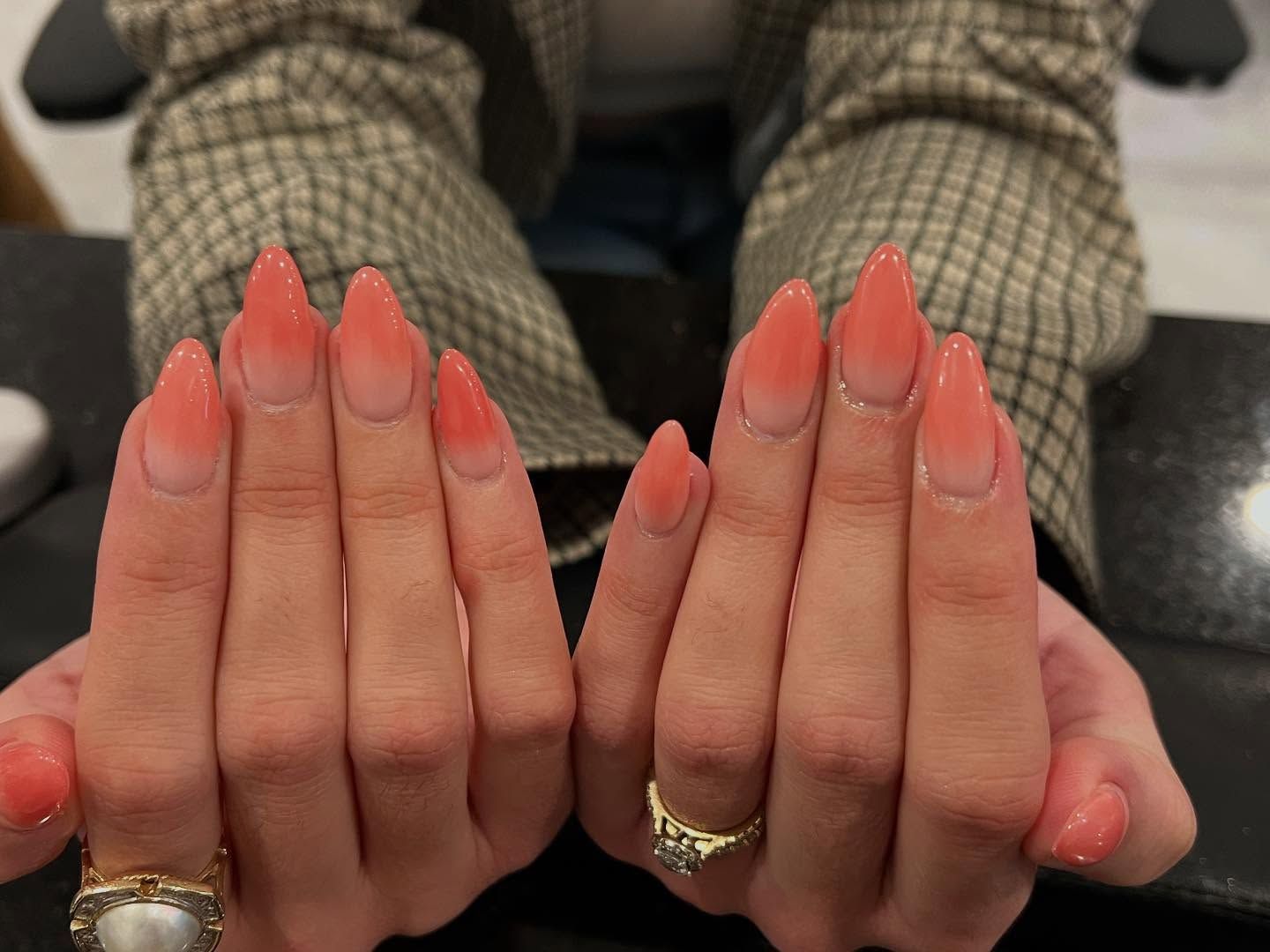 Hands with coral ombre stiletto nails resting on a surface, wearing a gold ring.