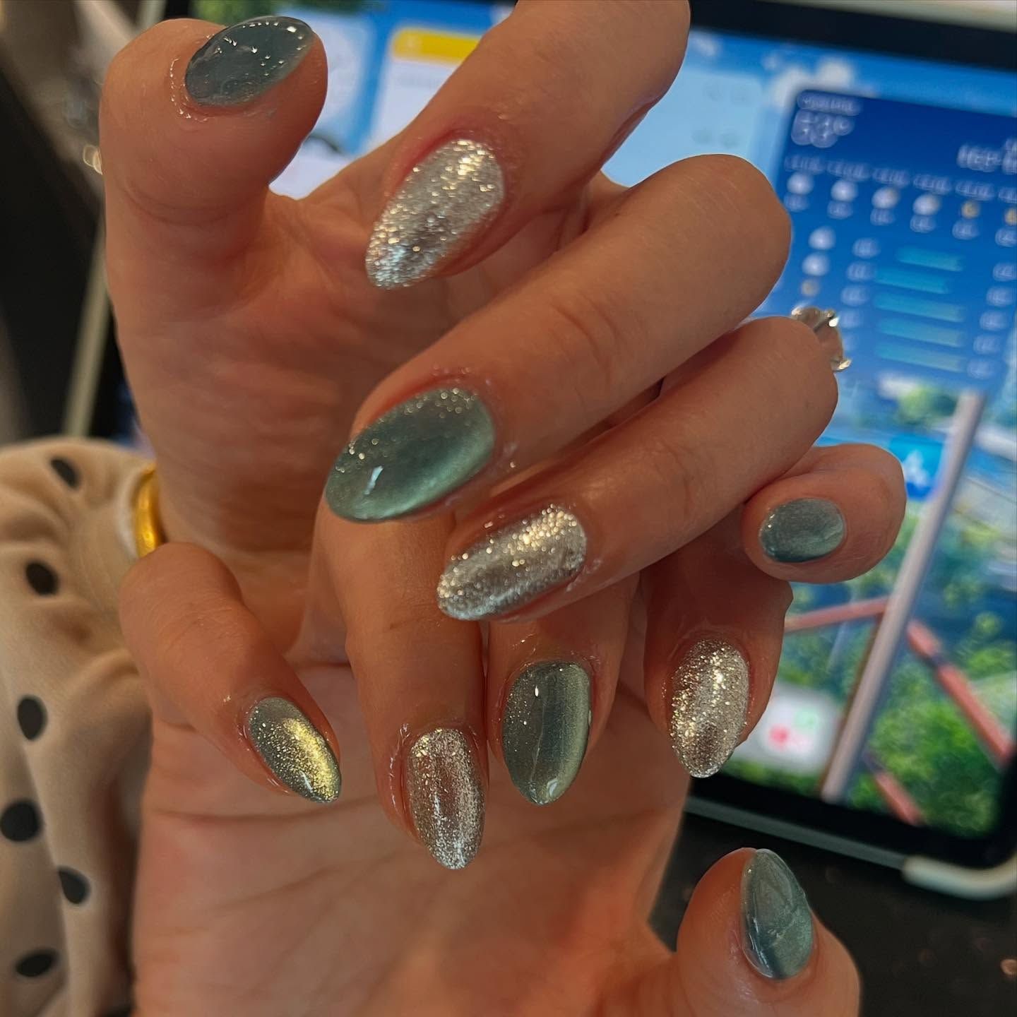 Hands with oval-shaped nails, some silver glitter, some sage green with cat-eye effect, on a blurred background.