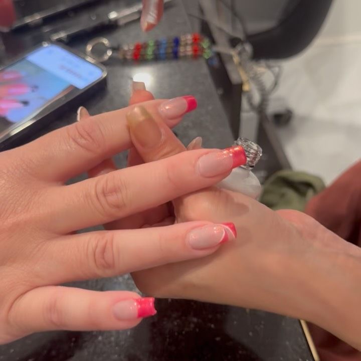 Hands with pink and white French tip nails touching. A phone and accessories in the background.