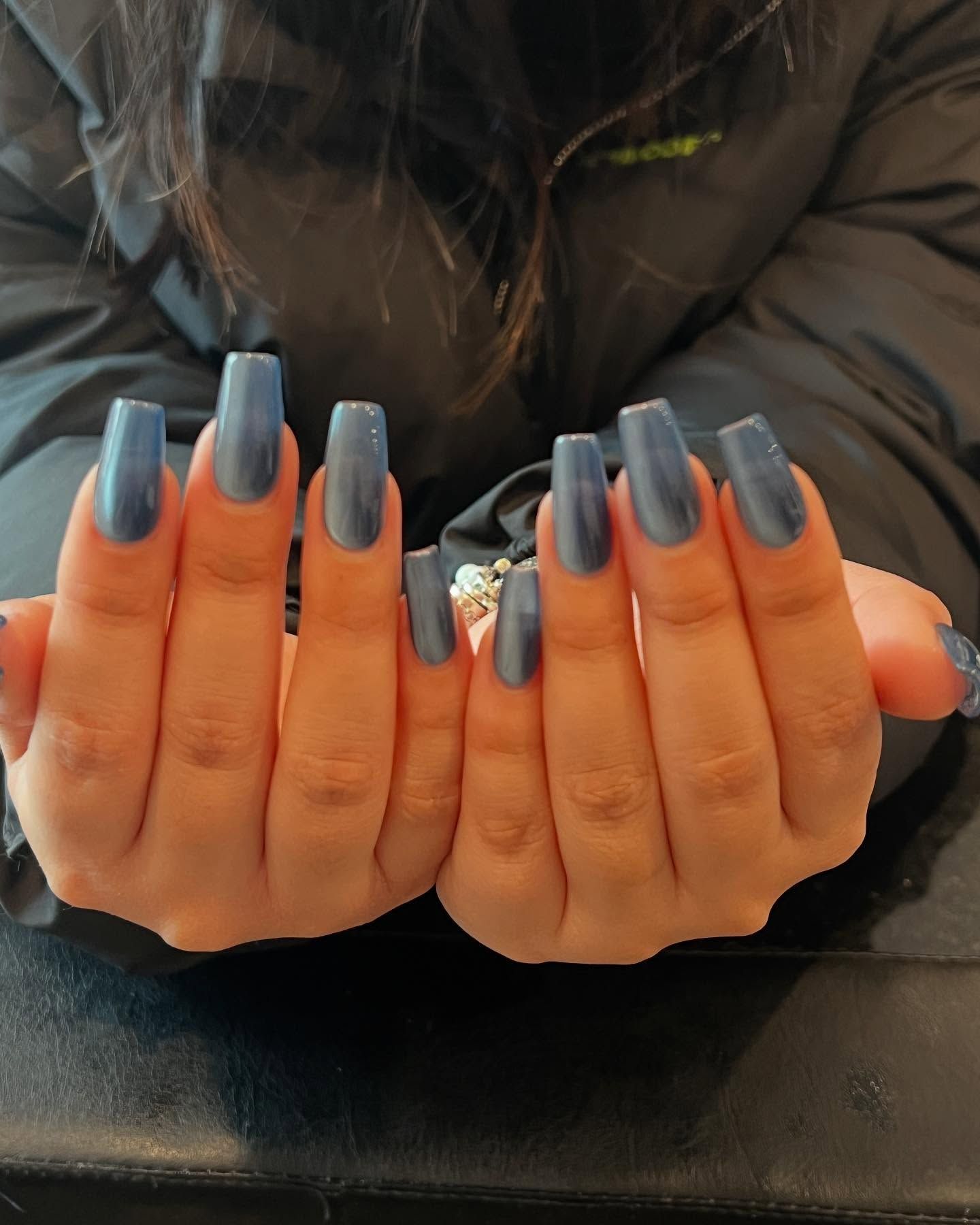 Hands with long, square-shaped blue nails. The background appears to be a dark-colored jacket.