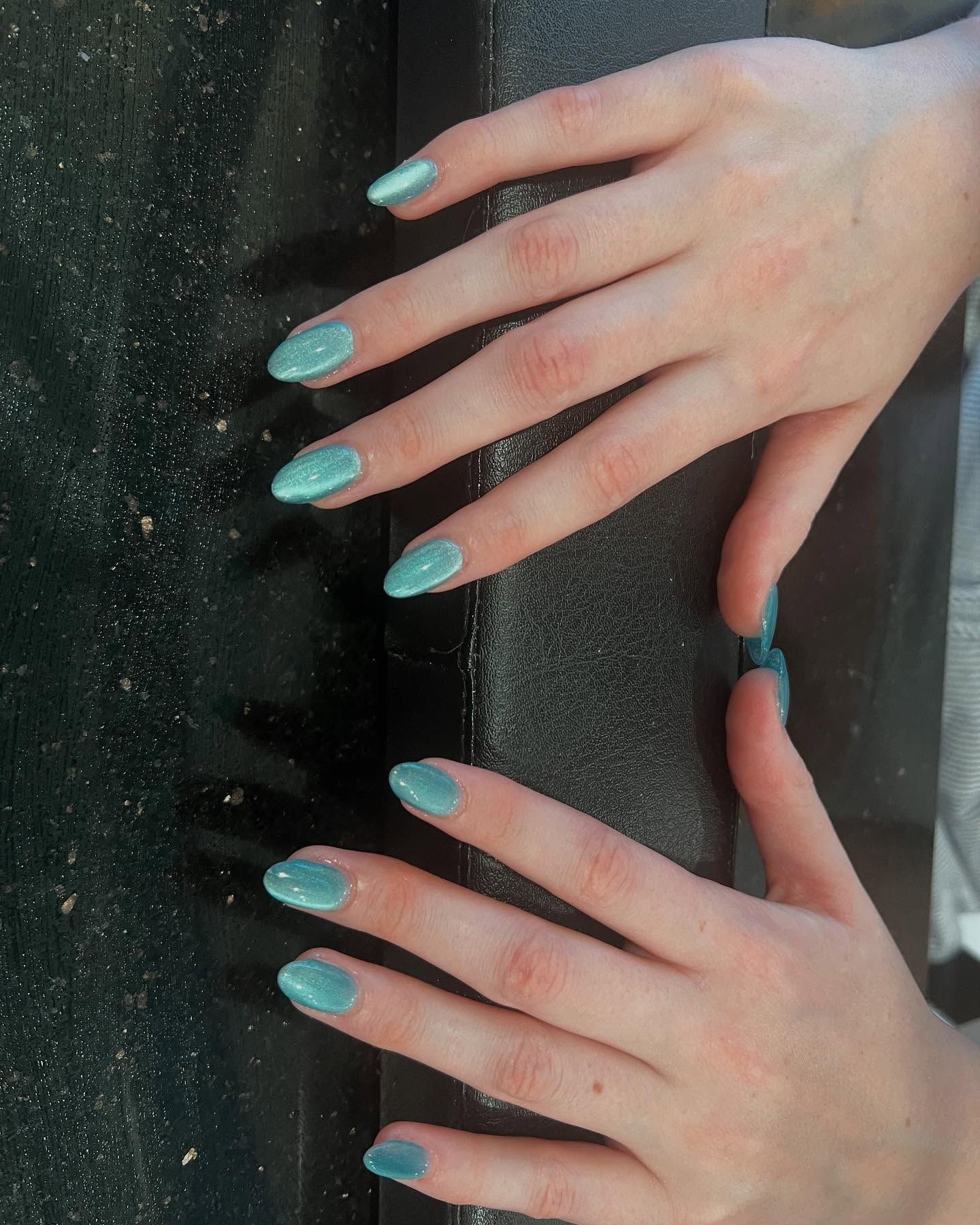 Hands with almond-shaped nails painted light blue with glitter, resting on a dark surface.