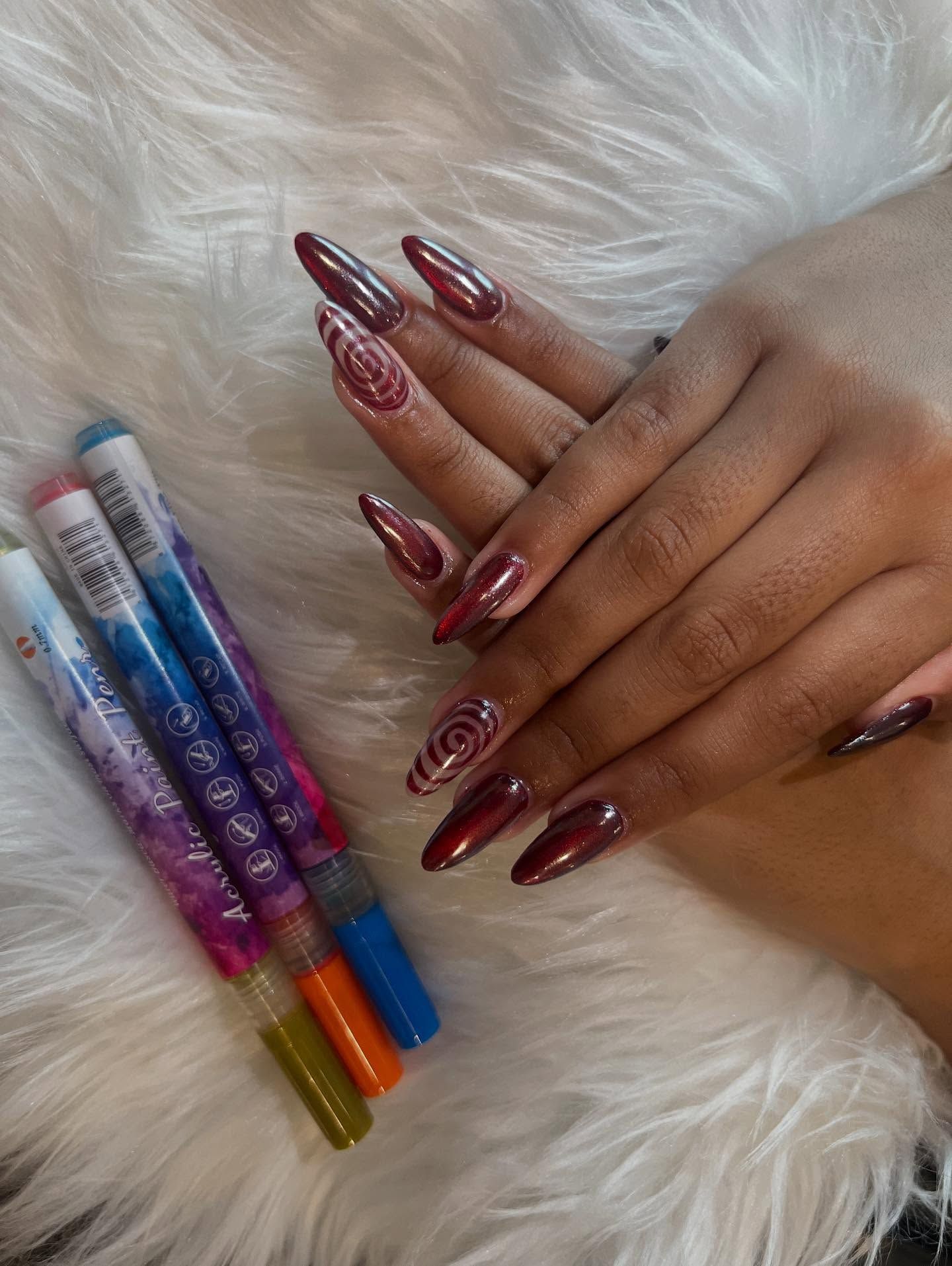 Long, burgundy nails with spiral art, held over a white, fluffy surface. Nail art pens alongside.