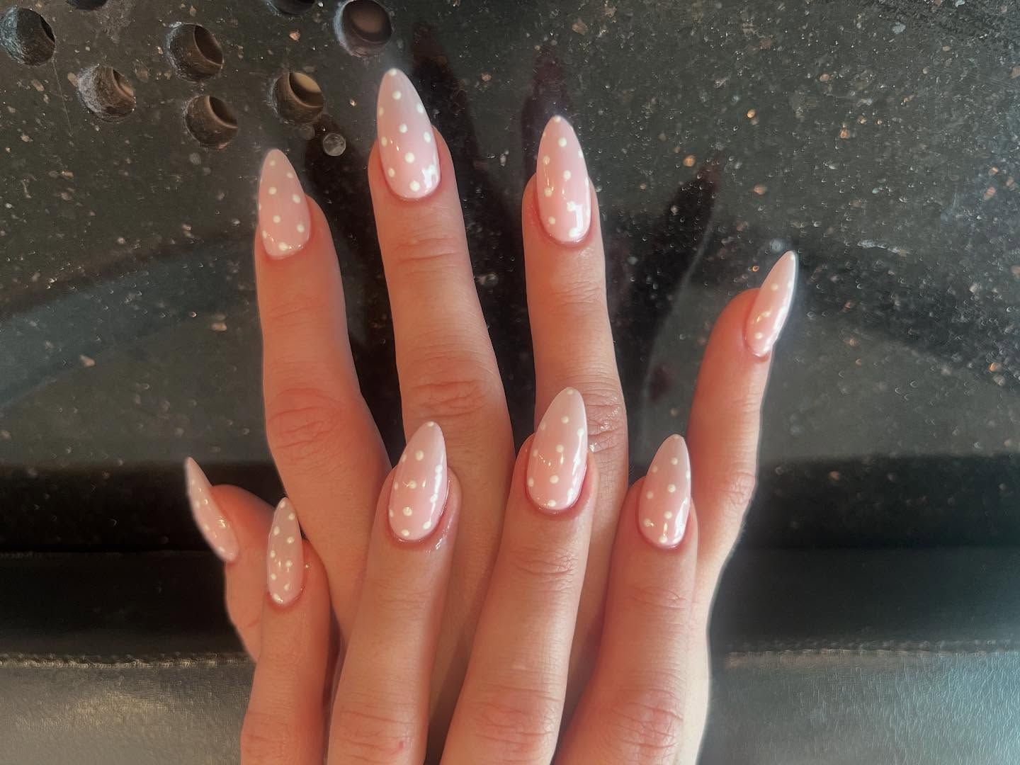 Hands with long, almond-shaped nails painted light pink with white polka dots.