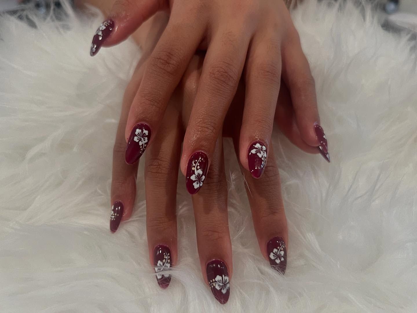 Burgundy-painted nails with white flower designs, on a white fluffy surface.