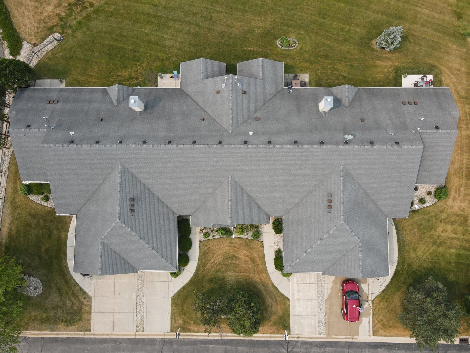 Aerial view of a gray-roofed duplex with a red car parked in a driveway on a grassy lot.