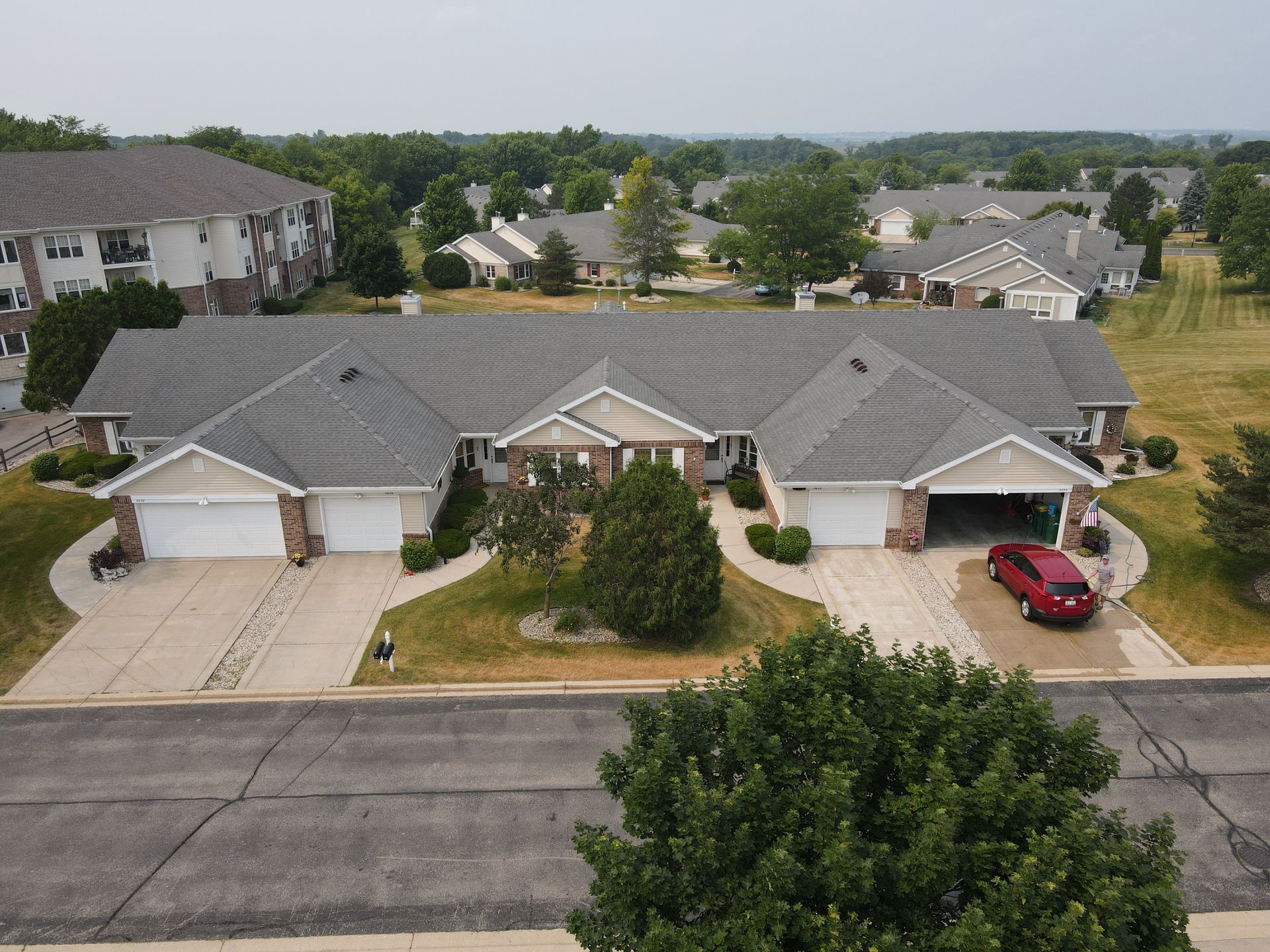 Townhouses with gray roofs, garages, and red car in driveway. Green trees and grass surround the buildings.